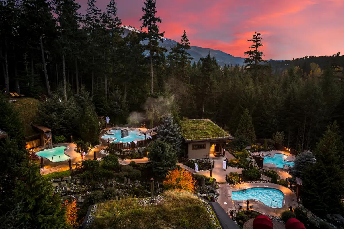 Aerial view of Scandinave Spa Whistler at dusk with glowing thermal pools and forest garden beneath sunset sky.