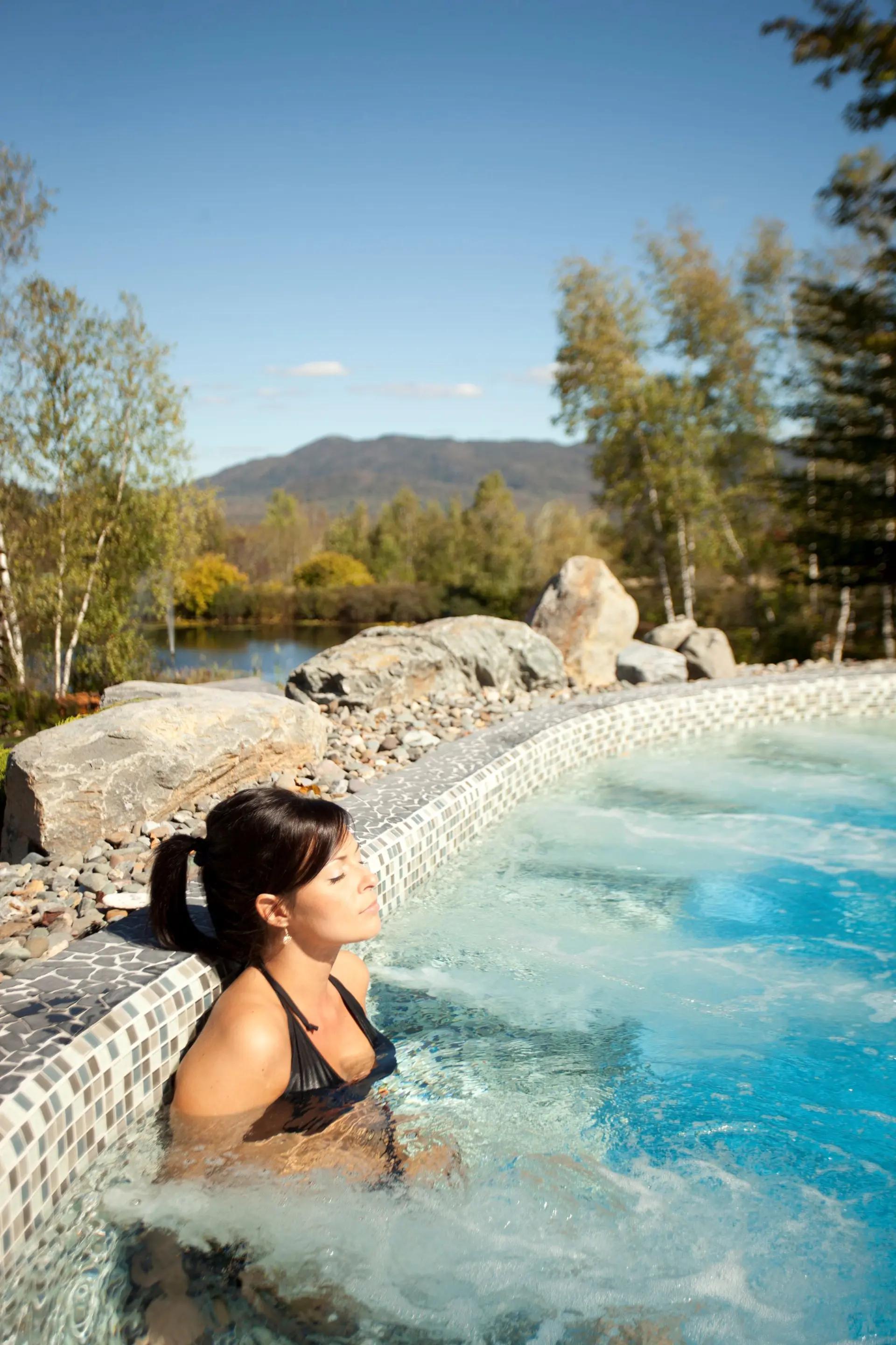 A woman relaxes with eyes closed in an outdoor thermal bath at Spa Eastman, surrounded by natural boulders, birch trees, and a stunning mountain and lake backdrop.