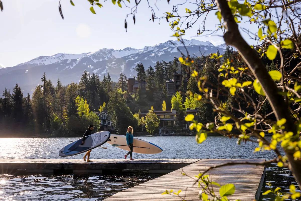 Two paddleboarders on a dock at a Whistler lake with Whistler and Blackcomb mountains in the background.