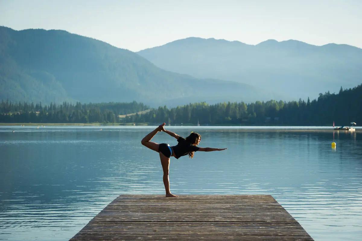 Woman in dancer's yoga pose on a dock overlooking a calm Whistler lake with forested mountains reflected in still water.