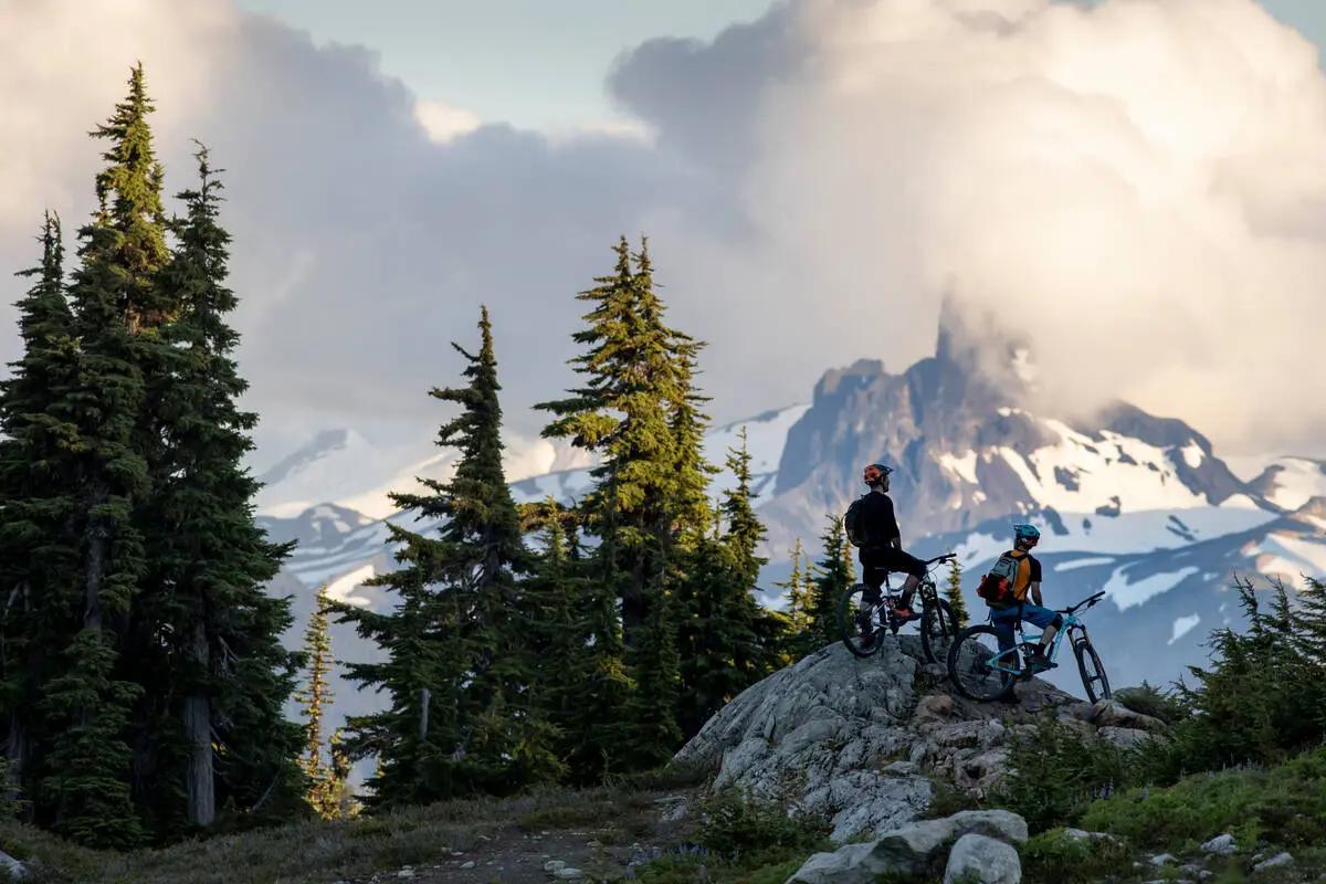 Two mountain bikers on a rocky alpine outcrop in Whistler with Coast Mountain peaks and subalpine forest.