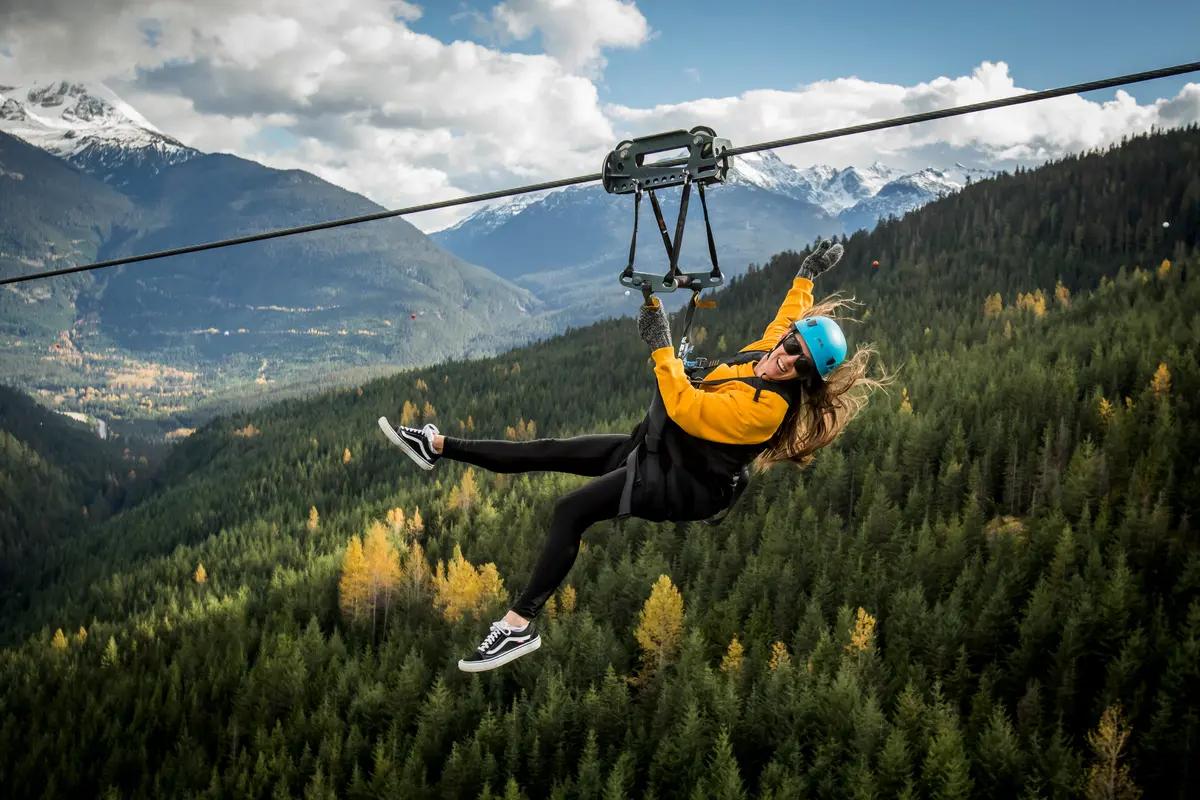 Woman on a mountain-to-mountain zipline above Whistler's forested valley with snow-capped Coast Mountain peaks.