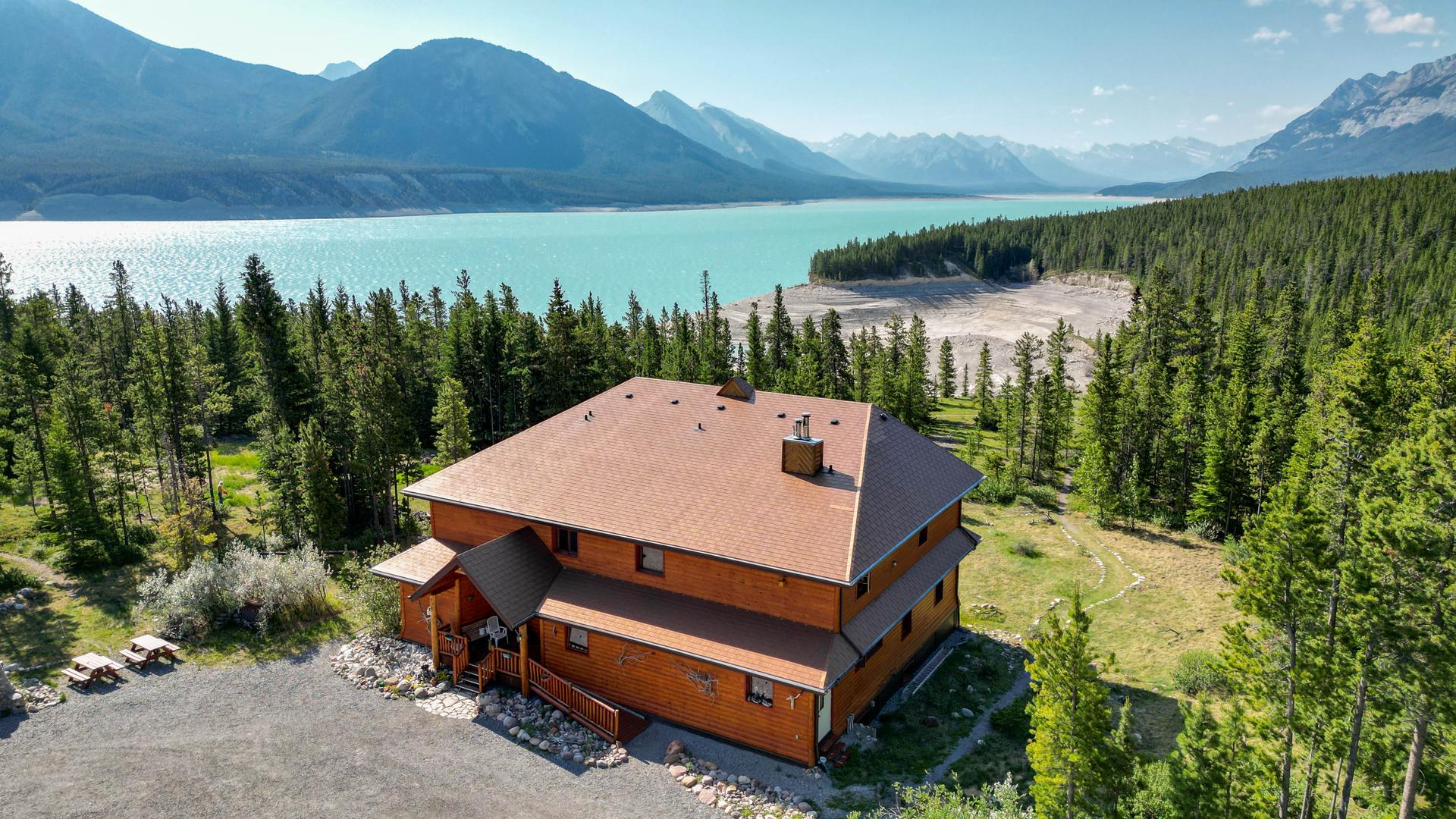 Aerial view of Aurum Lodge, a two-storey log-sided building surrounded by evergreen forest on the north shore of turquoise Abraham Lake in Bighorn Country, Alberta, with the Rocky Mountains in the background