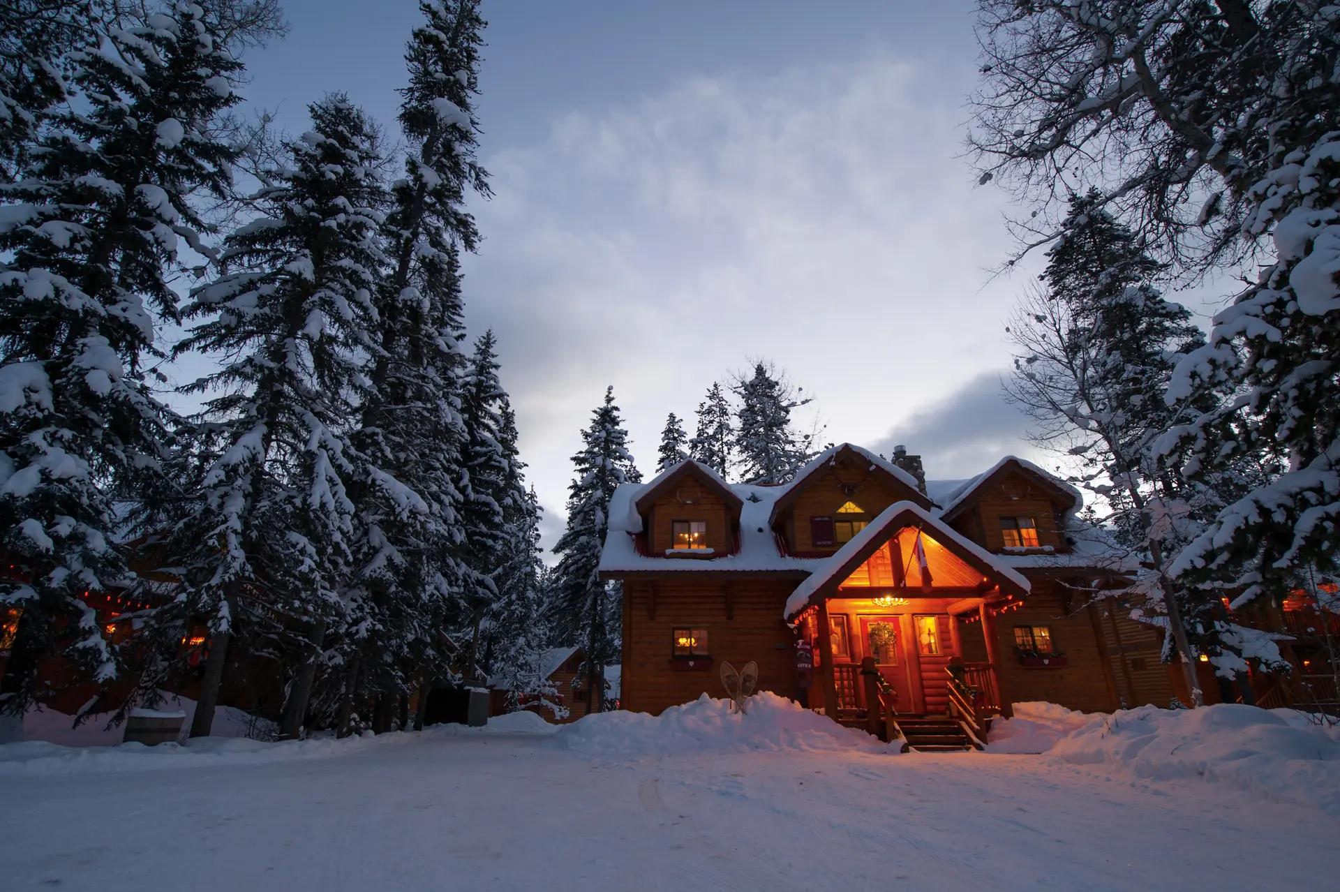 Warmly lit log cabin at Baker Creek Mountain Resort glowing at dusk, surrounded by snow-laden evergreens in Banff National Park, Alberta.