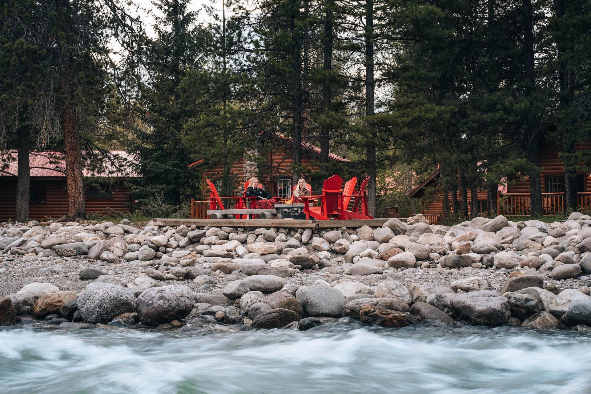 Two guests wrapped in blankets sitting in red Muskoka chairs on a riverside deck at Baker Creek Mountain Resort, with log cabins and pine forest behind them, Banff National Park, Alberta