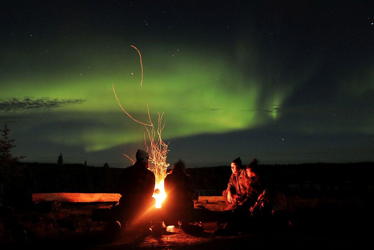 Guests gather around a campfire beneath a bright green aurora display at a remote lodge in Canada's Northwest Territories.