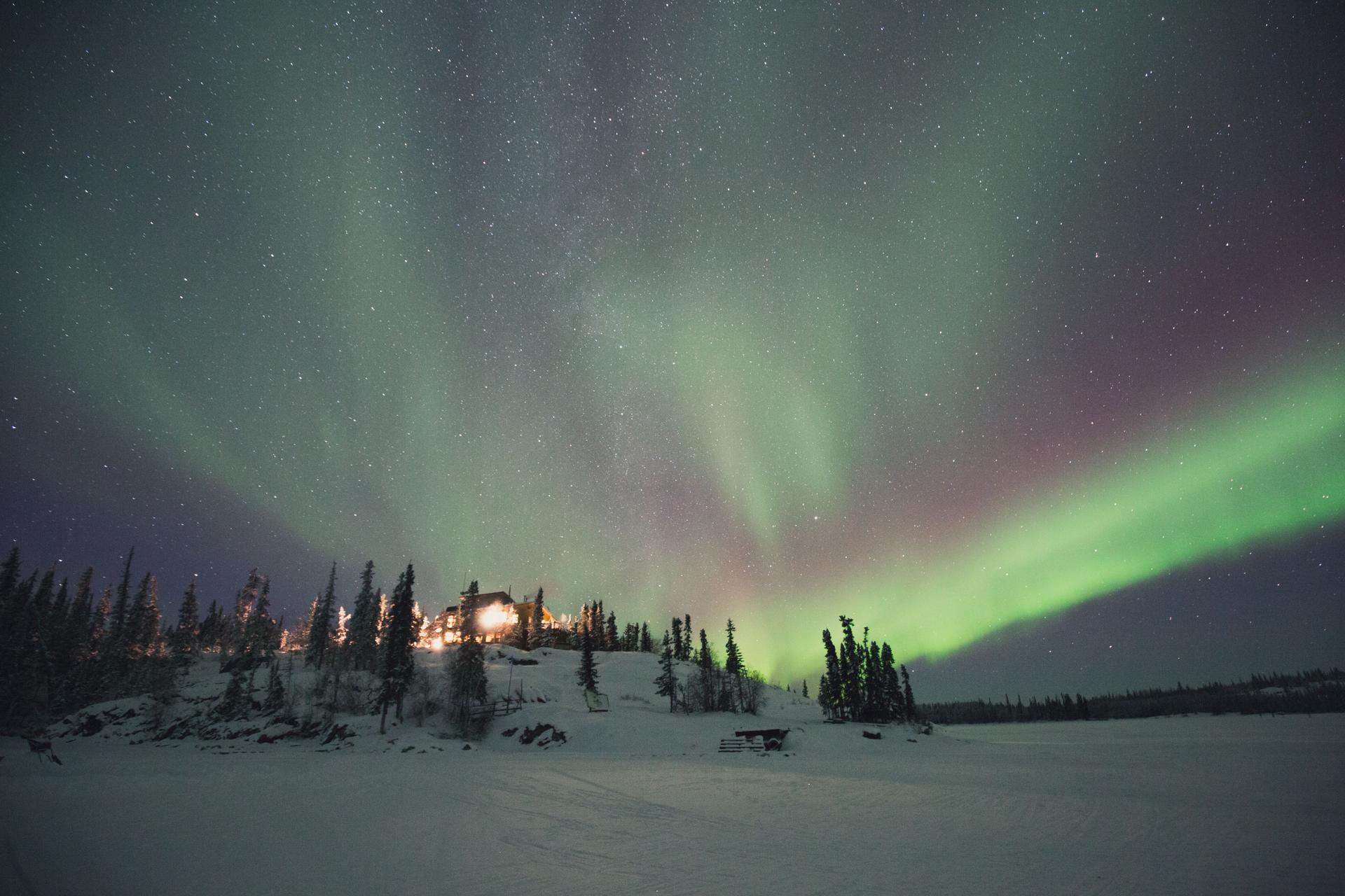 A wide view of green and purple aurora borealis streaking across the sky above the illuminated Blachford Lake Lodge on a snowy, tree-lined shore.