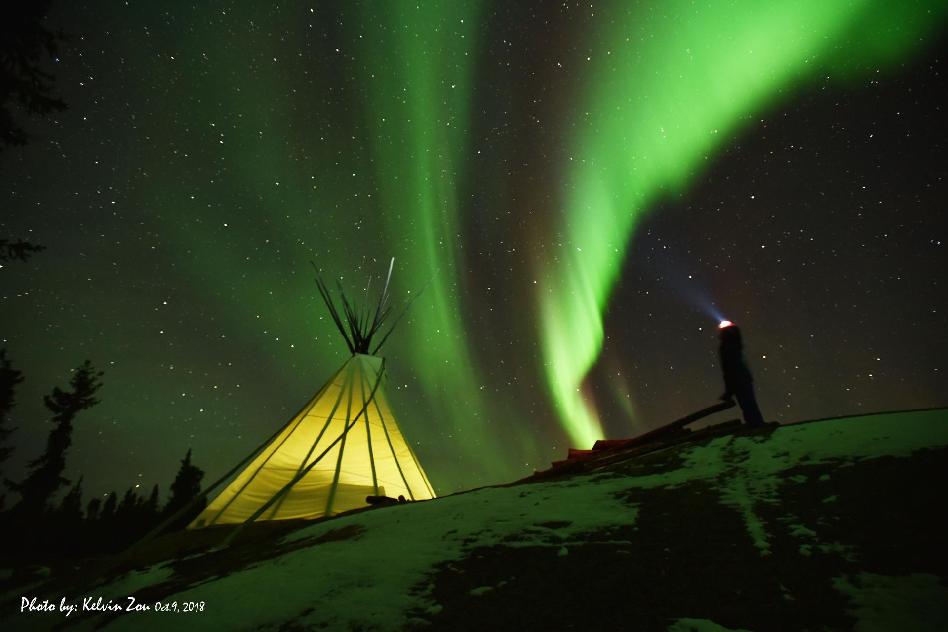A glowing teepee lit from within stands on a snowy ridge as a person shines a headlamp upward beneath a bright green aurora display.
