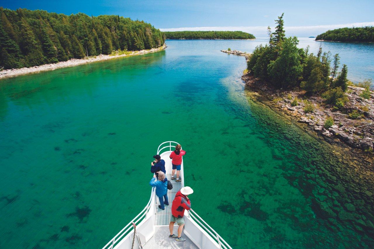 People stand on the bow of a tour boat cruising through the clear turquoise waters of Bruce Peninsula, surrounded by forested islands
