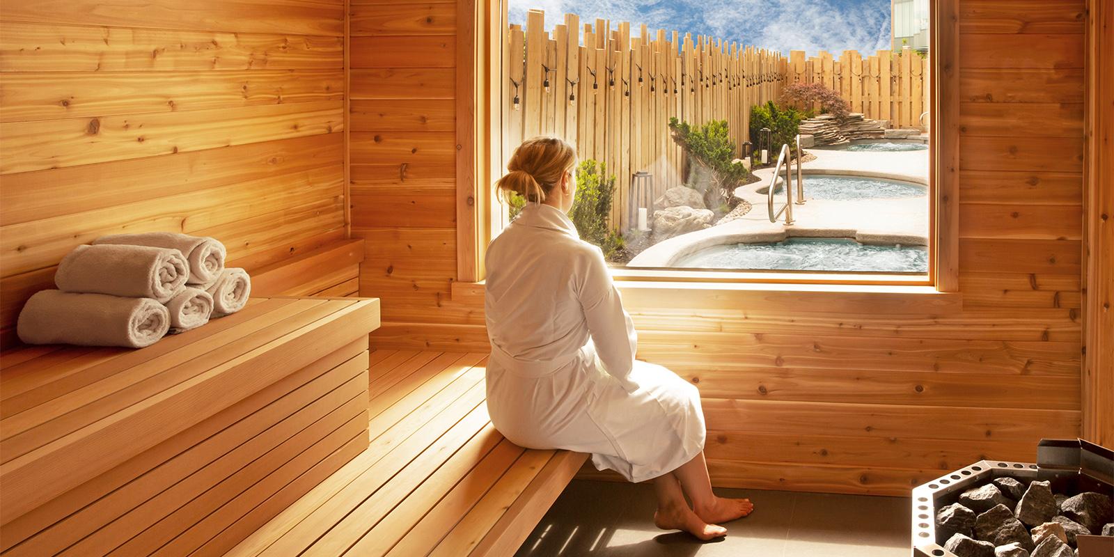 Guest in white robe sitting inside cedar Finnish-inspired sauna cabin at Burning Springs Spa & Thermal Pools, with outdoor thermal pool circuit visible through window, Niagara Falls, Ontario
