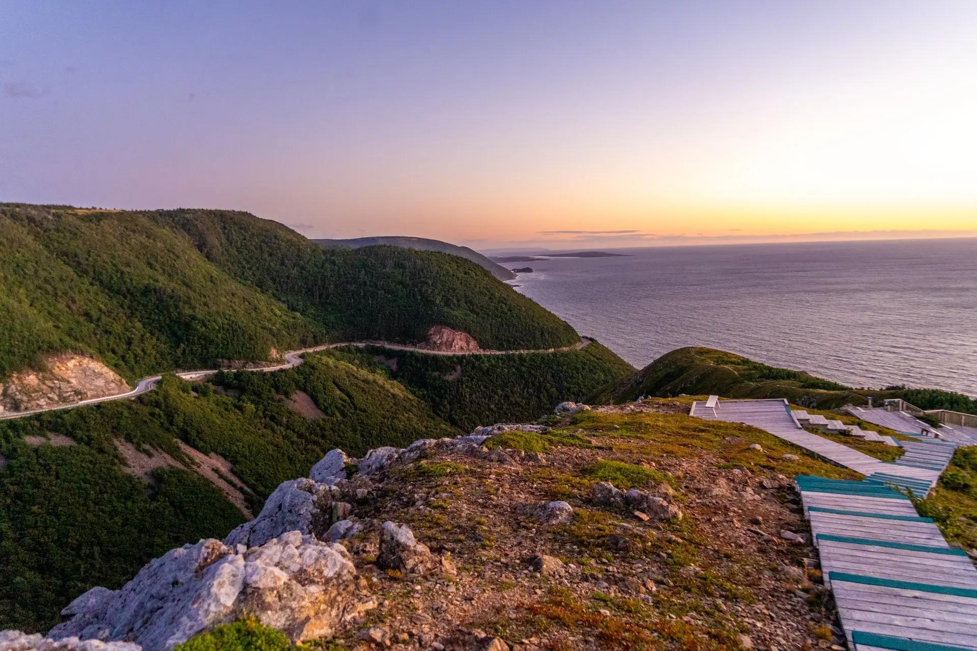 The wooden boardwalk of the Skyline Trail descends along clifftops in Cape Breton Highlands National Park, Nova Scotia, at sunset, with the Atlantic Ocean on the horizon.