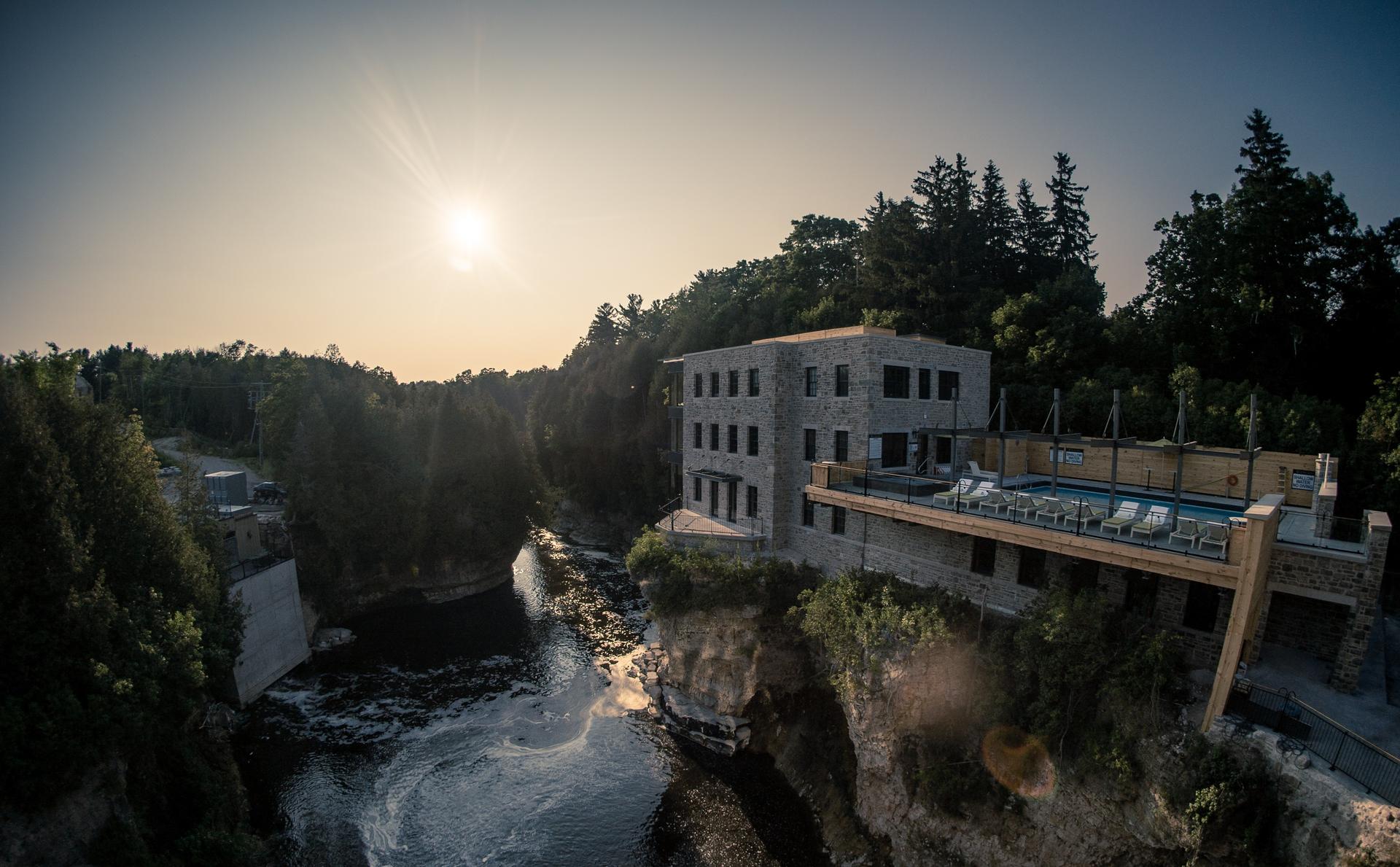 Aerial sunset view of Elora Mill Hotel & Spa perched on a limestone cliff above the Grand River gorge, with rooftop pool deck visible, Elora, Ontario