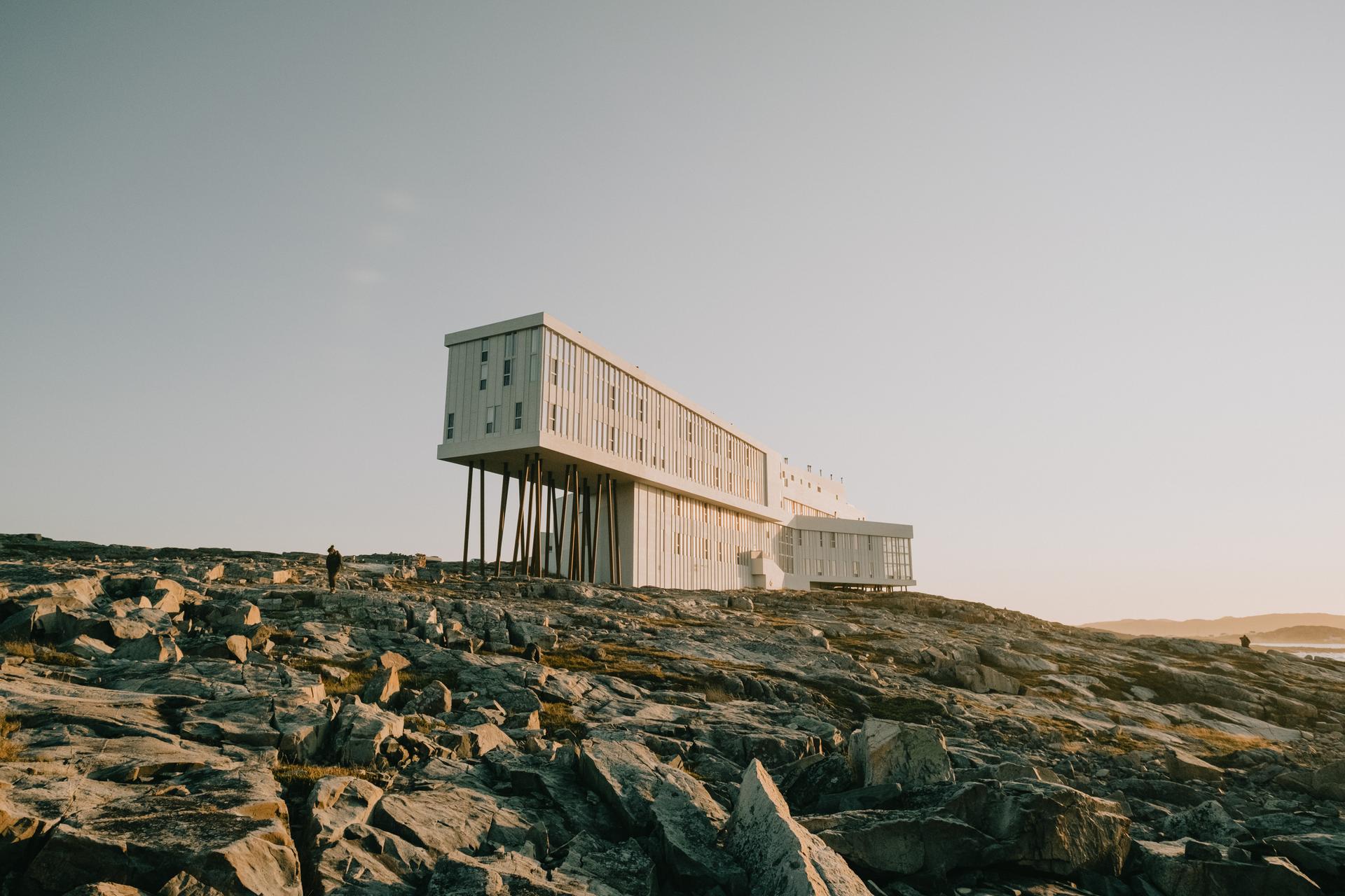 Fogo Island Inn's iconic elevated box structure perched on wooden stilts above the rocky shoreline of Fogo Island, Newfoundland, Canada, photographed at golden hour with a warm sunset sky.