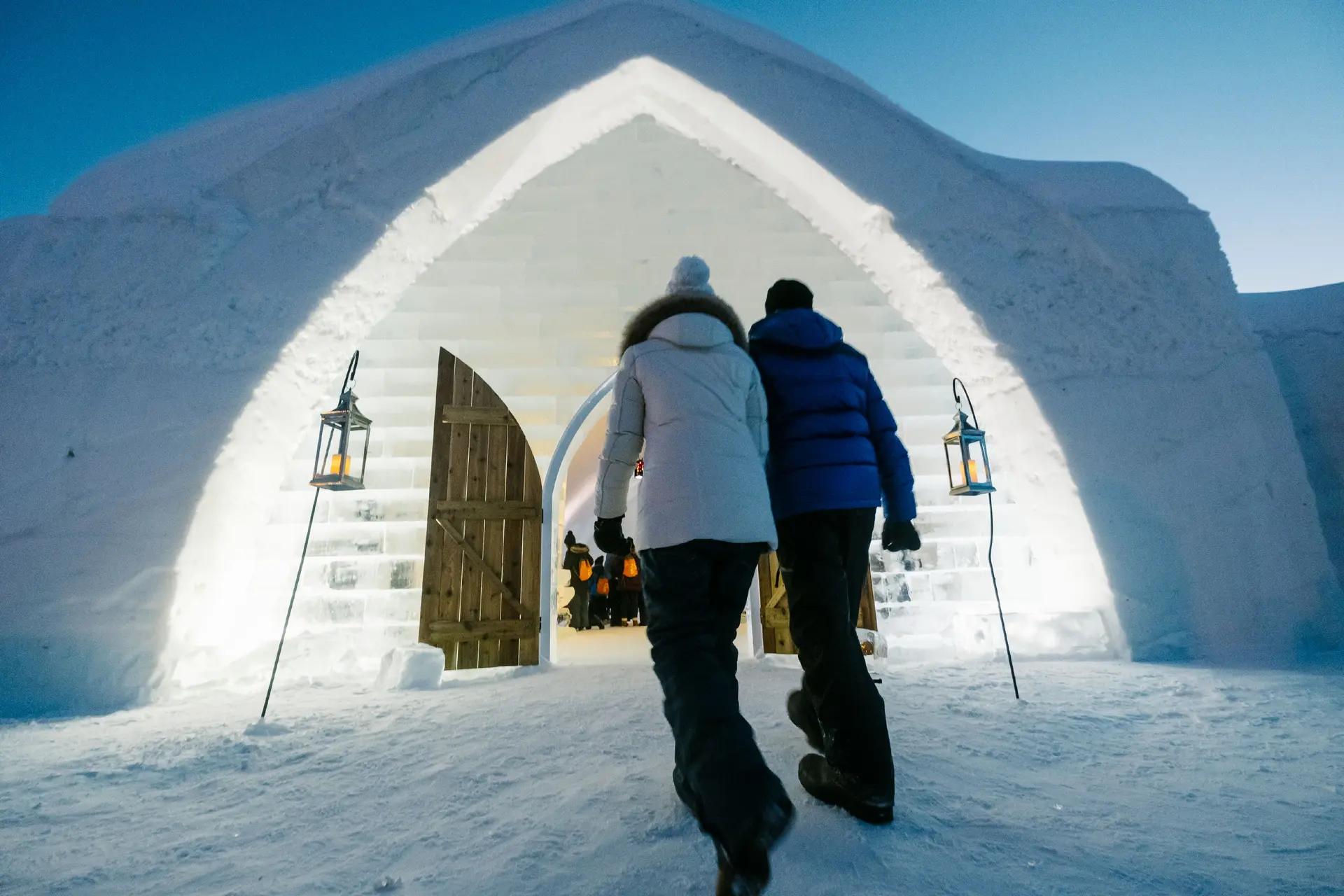 A couple walks through the illuminated arched entrance of Hôtel de Glace, a fortress-like structure built entirely from ice and snow.