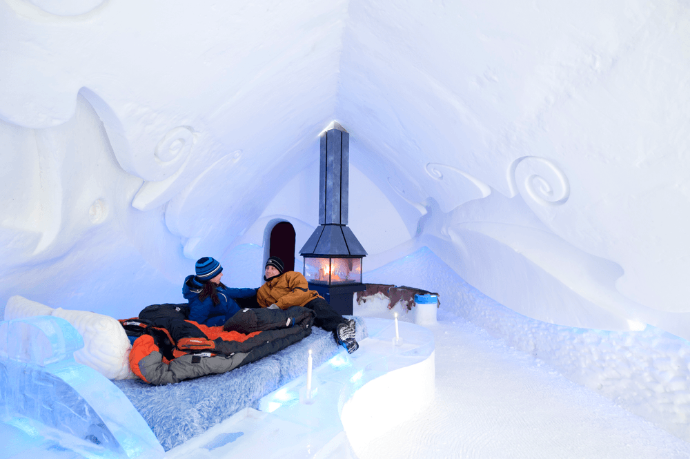 Two guests in sleeping bags relax on an ice bed beside a glowing fireplace inside Hôtel de Glace, Quebec City.