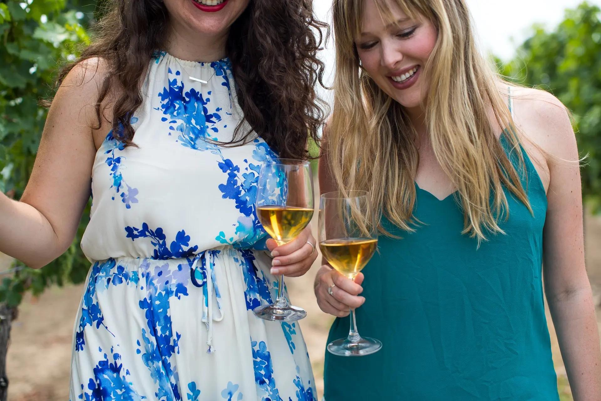 Two women hold glasses of white wine while walking through a vineyard.