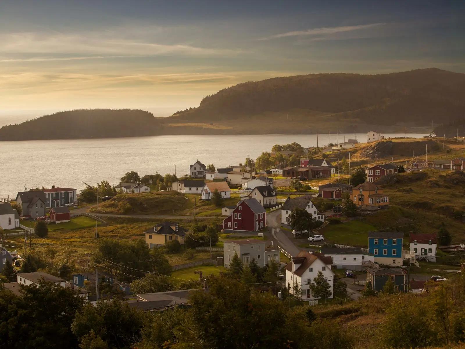 An aerial view of the colorful saltbox houses and winding roads of Port Rexton and Trinity in Newfoundland and Labrador, with a calm cove and rolling hills stretching toward the horizon.