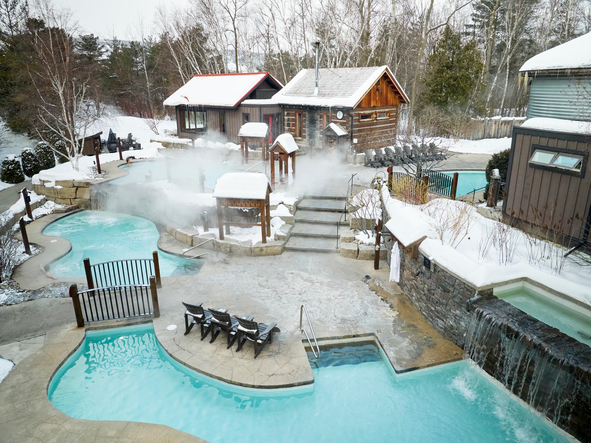Outdoor hot spring pools and cold plunge at Scandinave Spa Blue Mountain in winter, Collingwood, Ontario, with steam rising over snow-covered grounds and a log sauna cabin in the background
