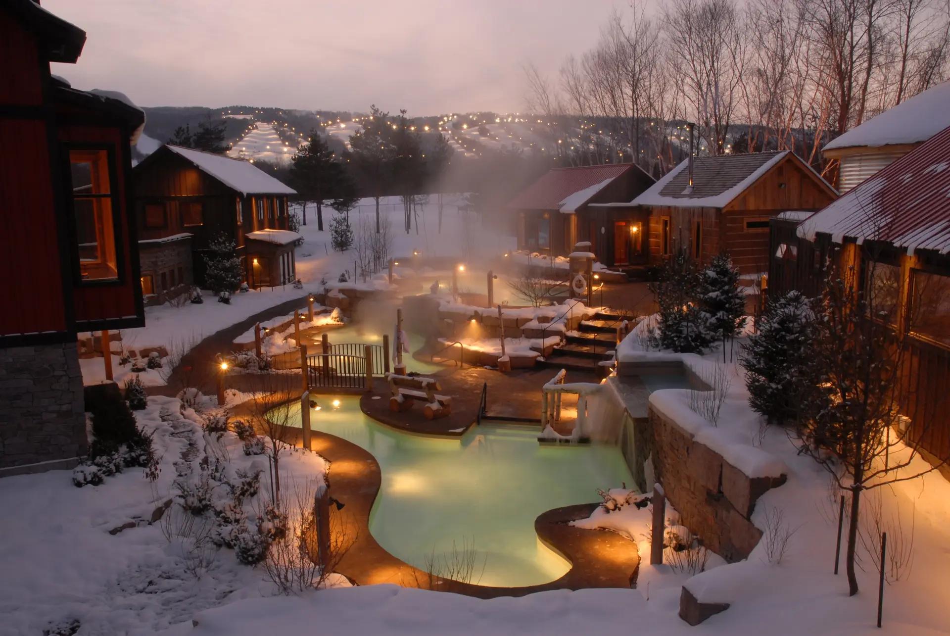 Aerial view of steaming outdoor thermal pools at Scandinave Spa Blue Mountain in winter, surrounded by snow-covered wooden cabins and warm pathway lighting, Collingwood, Ontario