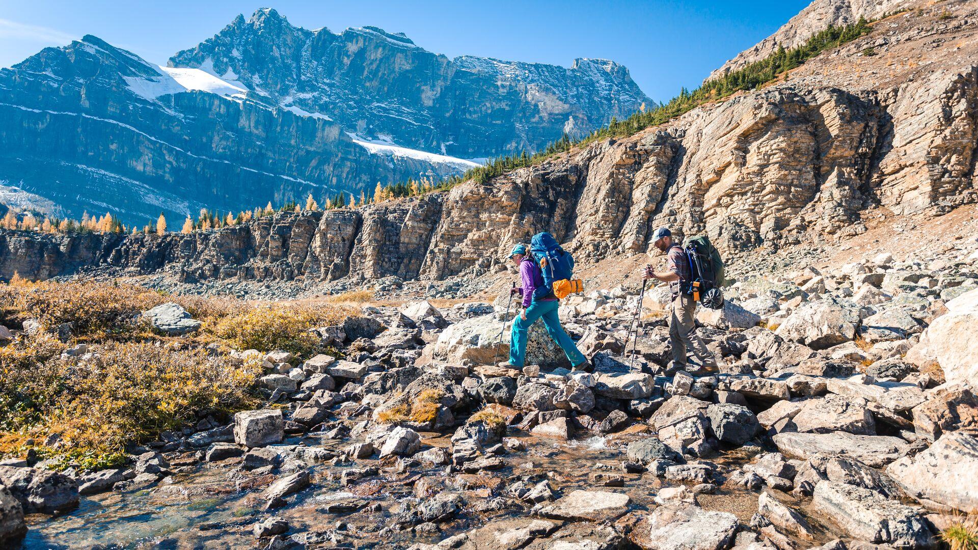 Two hikers with backpacks and trekking poles crossing rocky alpine terrain on the approach to Skoki Lodge, with golden larch trees and snow-capped peaks in Banff National Park, Alberta