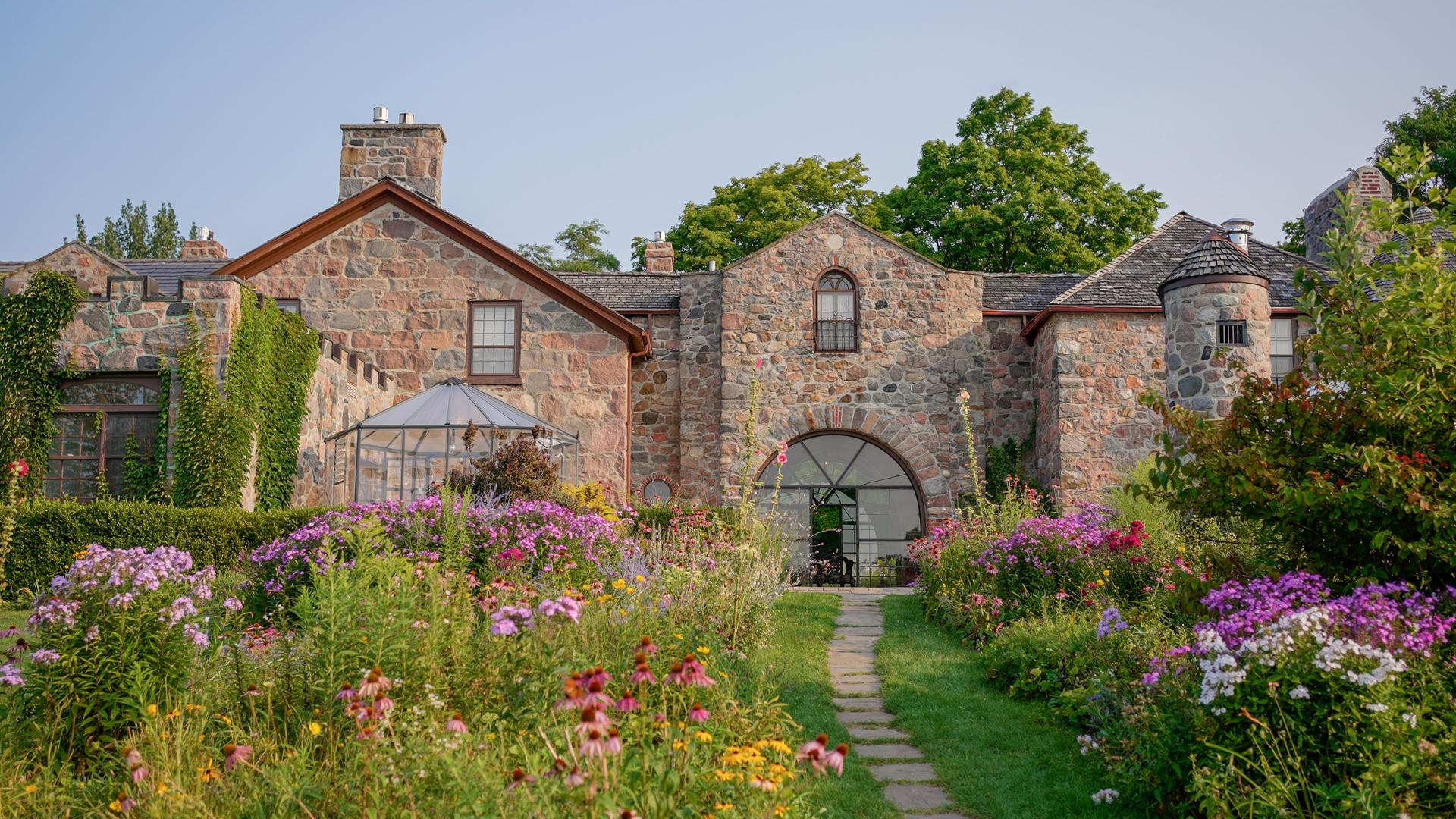 Wildflower garden path leading to the historic fieldstone buildings of Ste. Anne's Spa in Grafton, Ontario, with a glass greenhouse pavilion nestled among the blooms.
