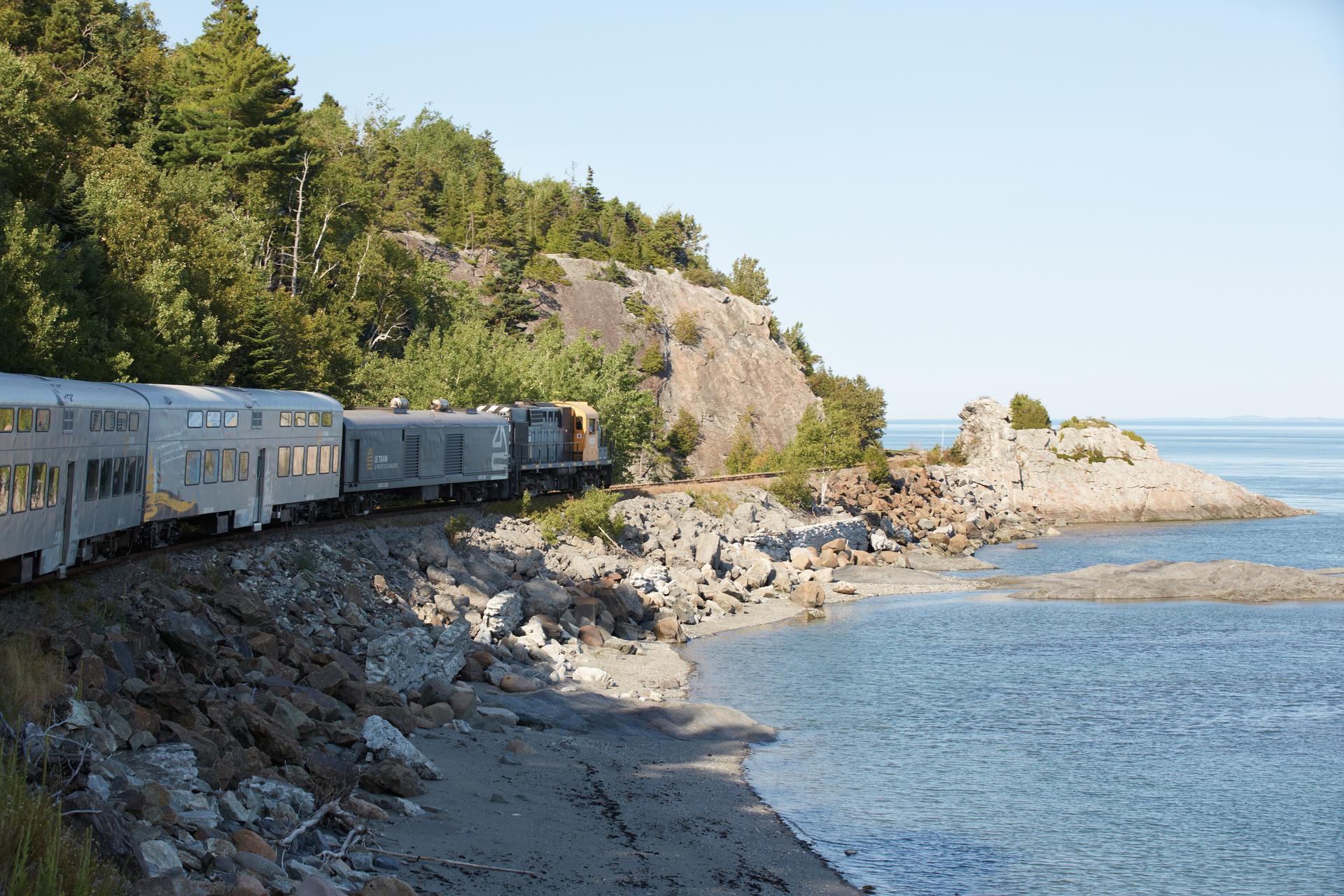 Train de Charlevoix passenger train curving along the rocky shoreline of the St. Lawrence River, surrounded by forested cliffs, on the scenic Québec City to La Malbaie route