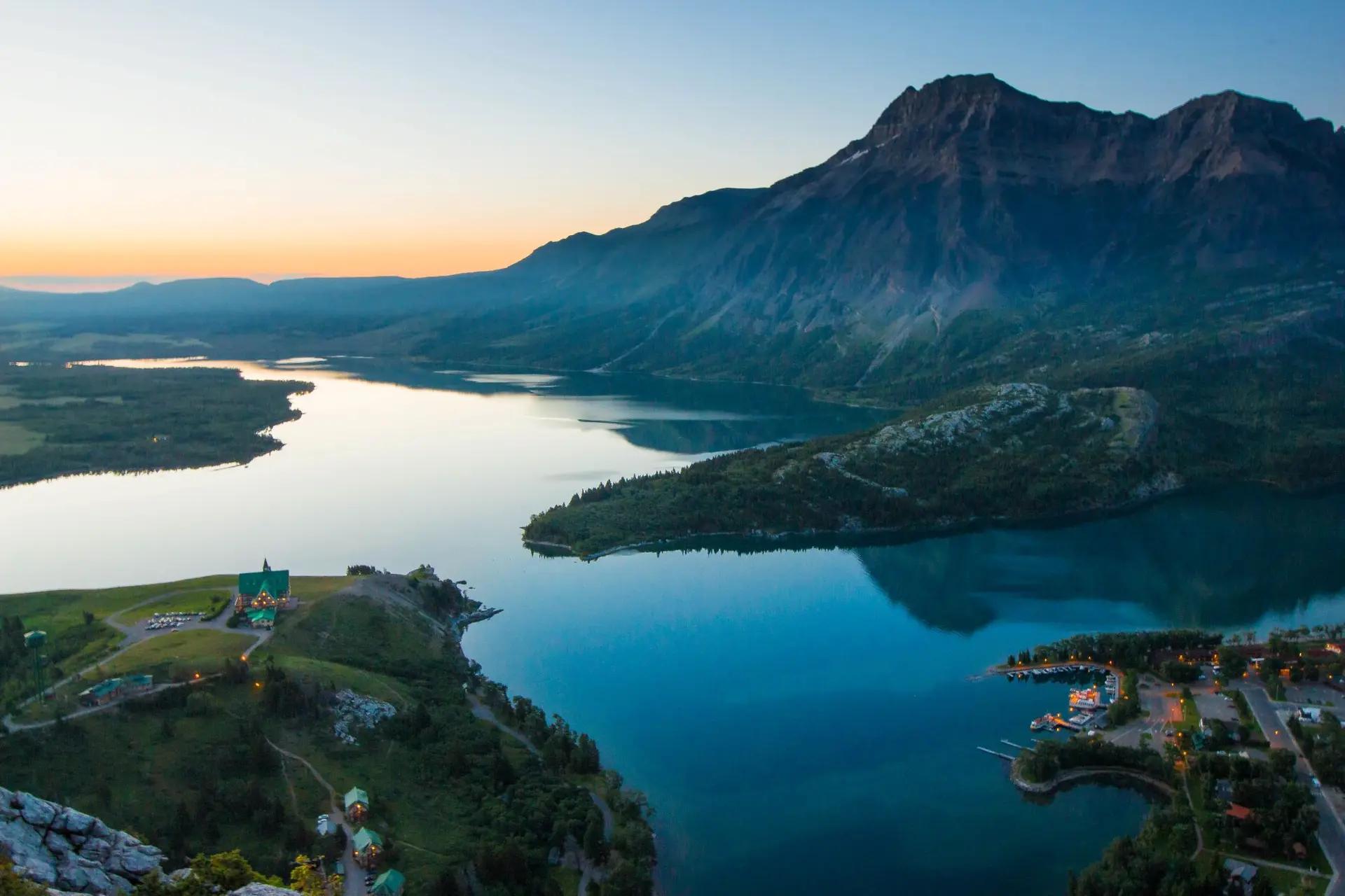 An aerial view of Waterton Lakes National Park in Alberta at dusk, with the Prince of Wales Hotel perched on a grassy bluff overlooking a lake with the Rocky Mountains in the background.