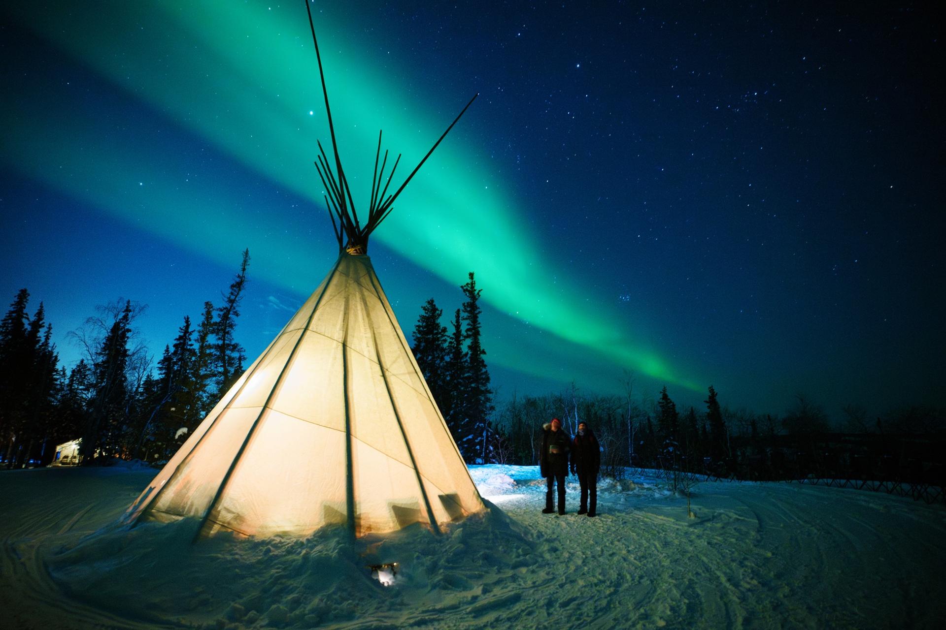 Two people standing beside an illuminated teepee beneath green northern lights
