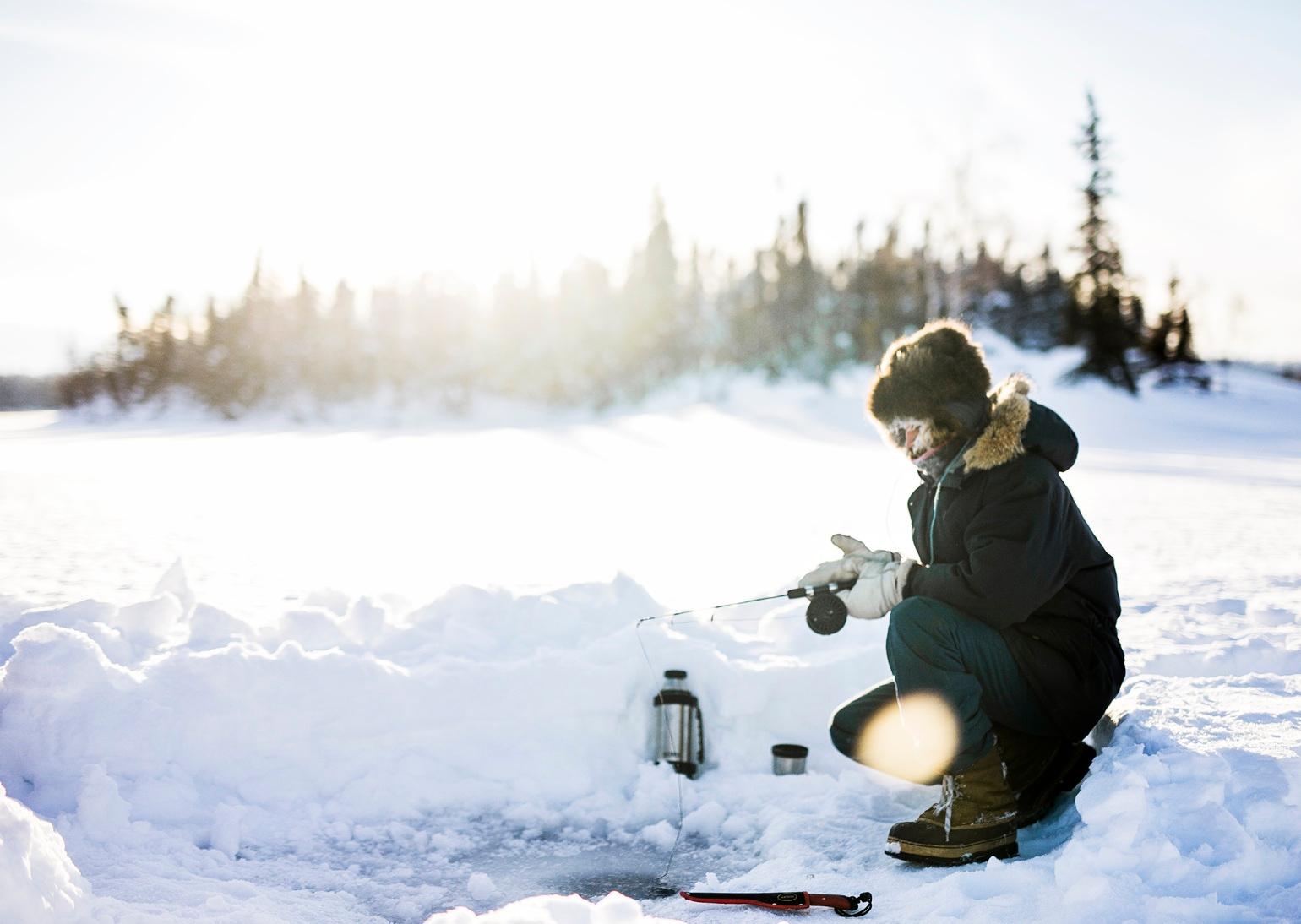 Person ice fishing on a snowy lake surrounded by evergreen trees