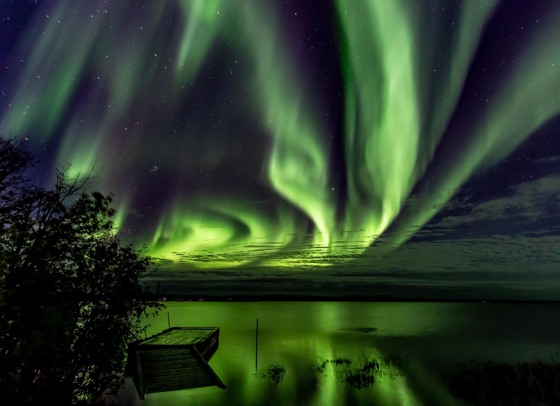 Vivid green northern lights reflecting on a calm lake beside a wooden dock