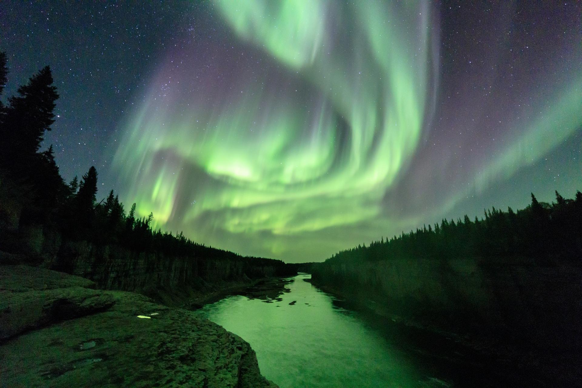 Green northern lights glowing above a forested river canyon under a starry sky