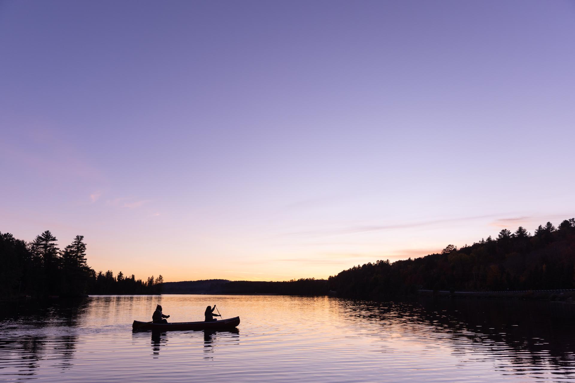 Two paddlers in silhouette canoe across a still lake at dusk near Killarney Lodge in Algonquin Provincial Park, with a pink and gold sunset reflecting on the water.