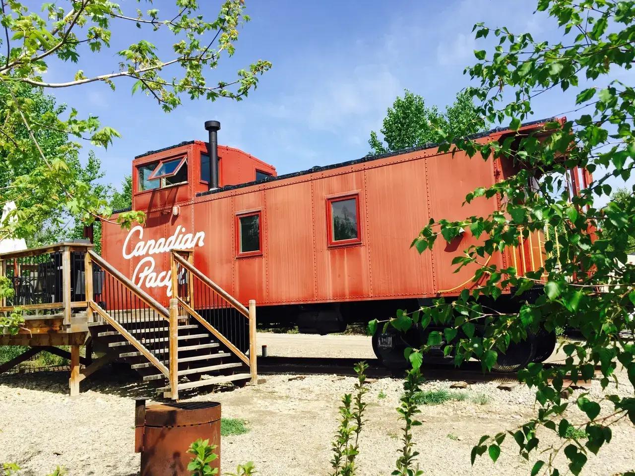 A restored Canadian Pacific red caboose railcar converted into a glamping cabin at Aspen Crossing near Mossleigh, Alberta, Canada, surrounded by trees on a sunny day