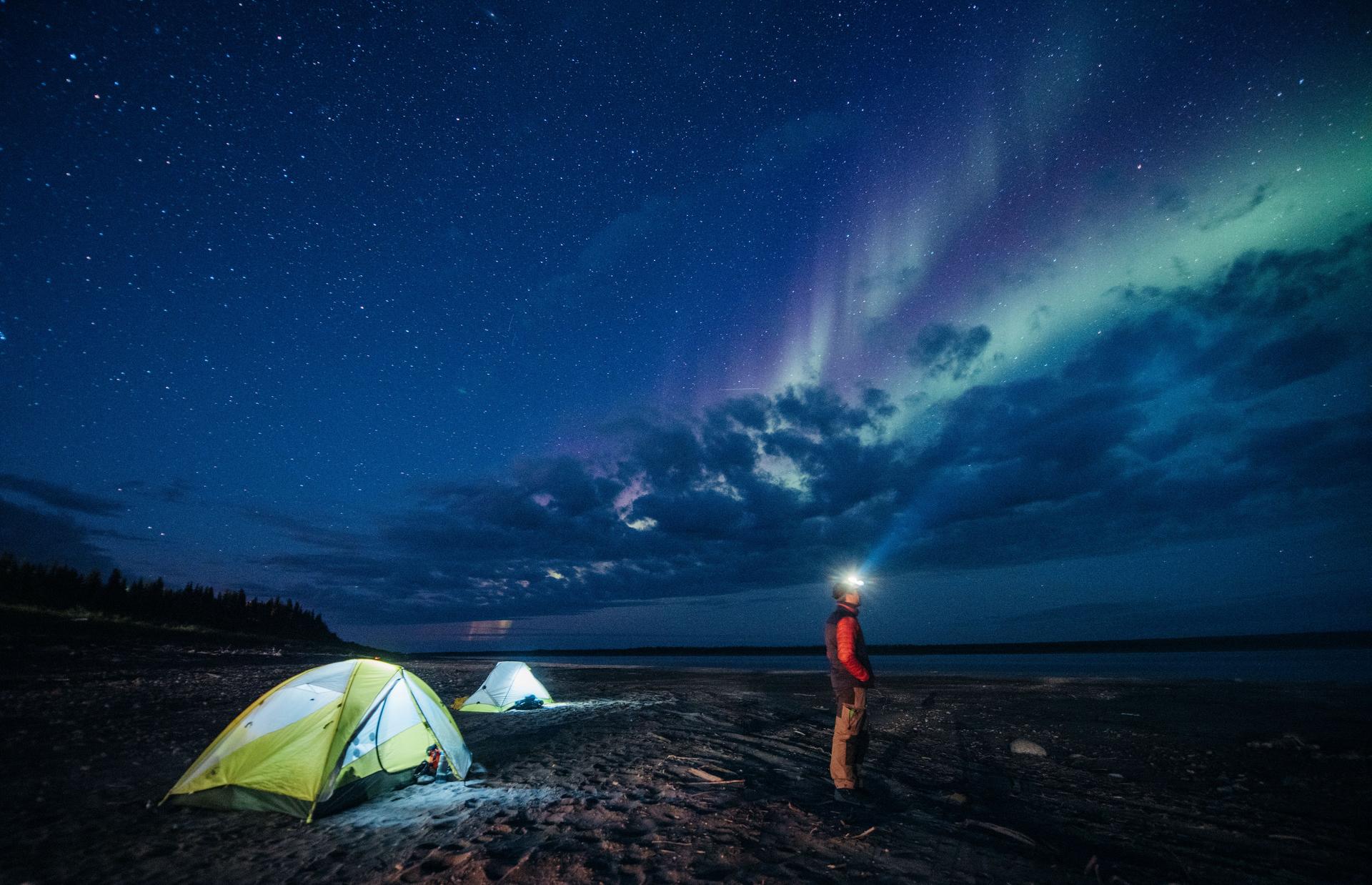 Camper wearing a headlamp watches the northern lights above two lit tents on a shoreline in the Northwest Territories