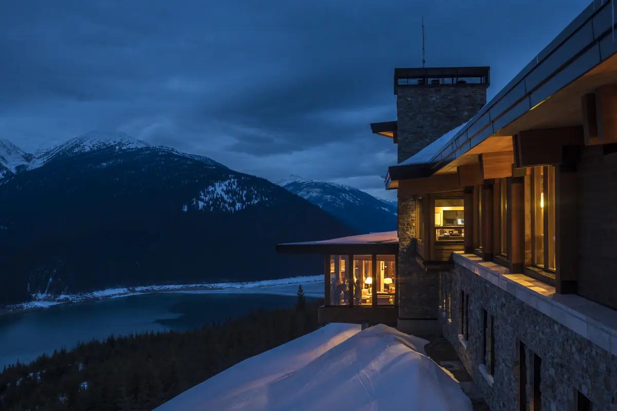 Warm light glows from inside Bison Lodge at dusk, perched above a snow-covered valley and dramatic mountain peaks in Revelstoke.