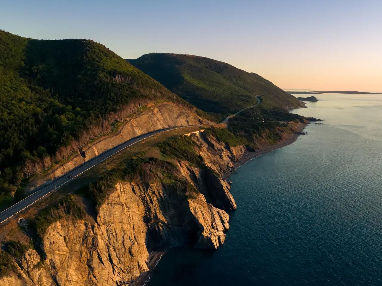 Aerial view of the Cabot Trail highway winding along dramatic golden-hued cliffs and forested mountain slopes above the Atlantic Ocean on Cape Breton Island, Nova Scotia, Canada, at sunset with a warm peach sky on the horizon