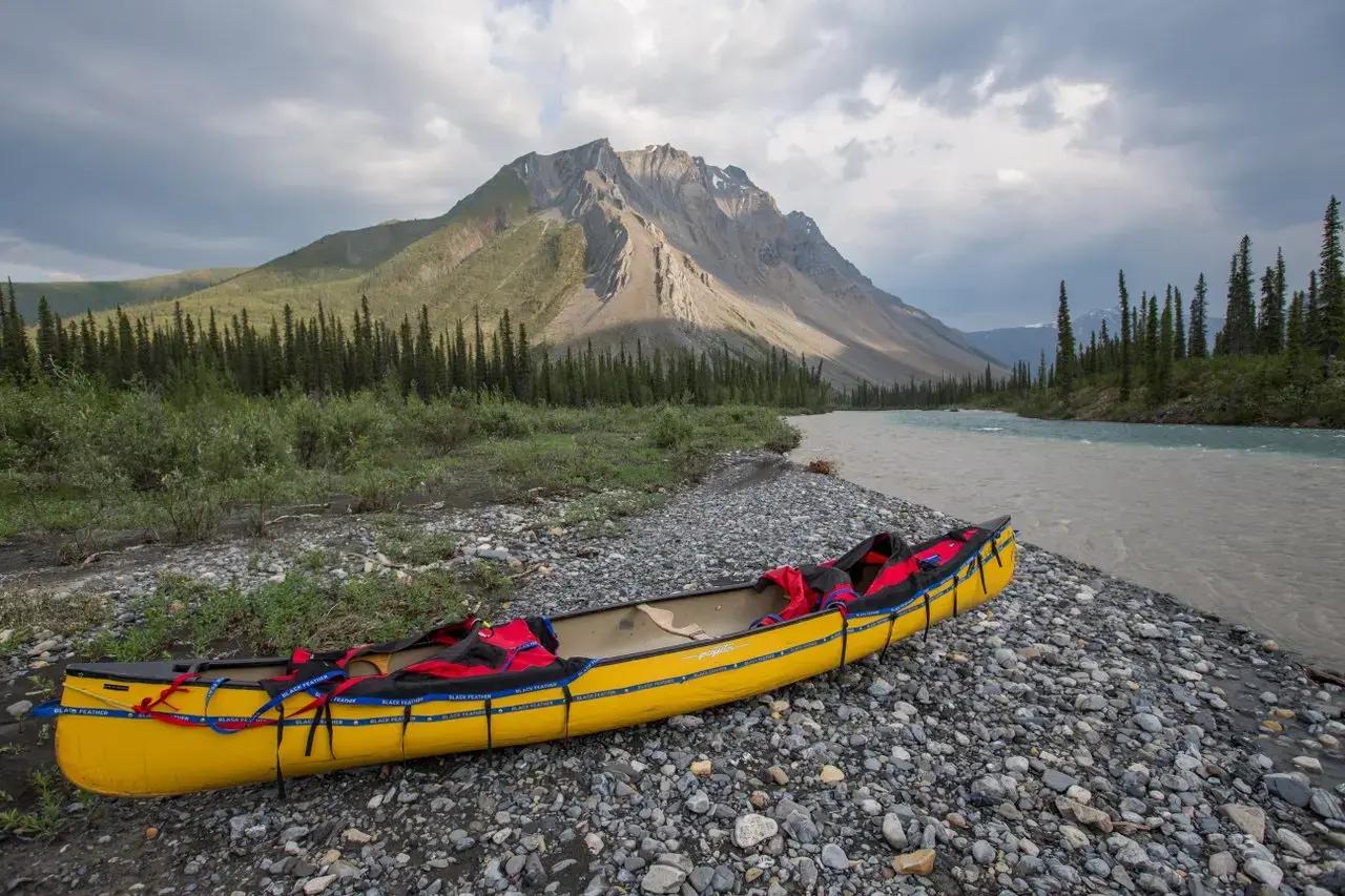 A yellow Black Feather inflatable canoe with red life jackets resting on a rocky gravel riverbank beside a glacial turquoise river with a dramatic rocky mountain peak and dense boreal spruce forest in the background in the Northwest Territories, Canada