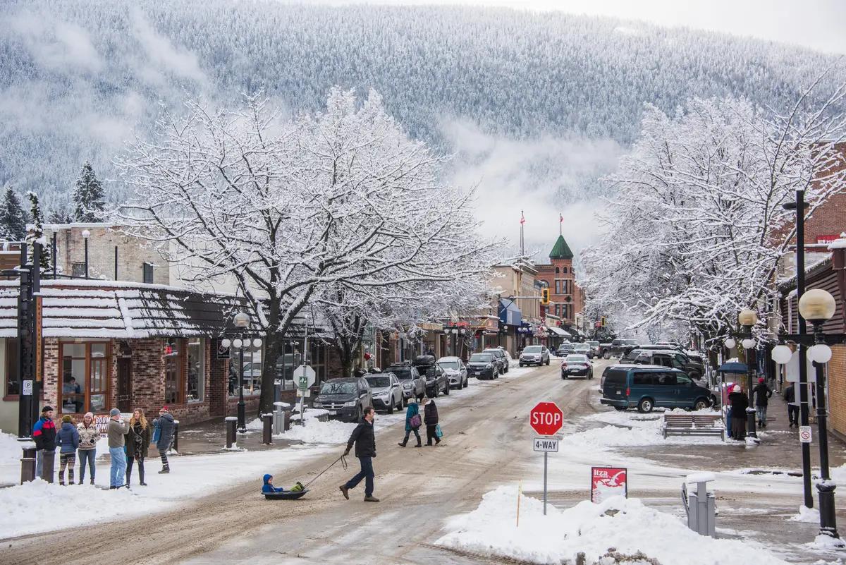 Pedestrians and snow-dusted trees line Nelson's historic main street in winter, with forested mountains rising dramatically in the background.