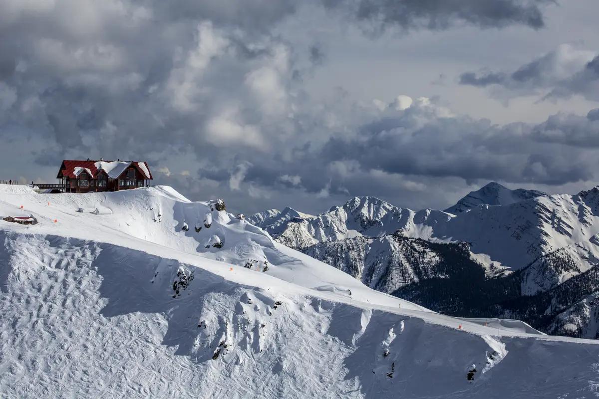 A red-roofed mountain lodge sits perched at the summit of Kicking Horse Mountain Resort, surrounded by vast snow-covered Rocky Mountain peaks.