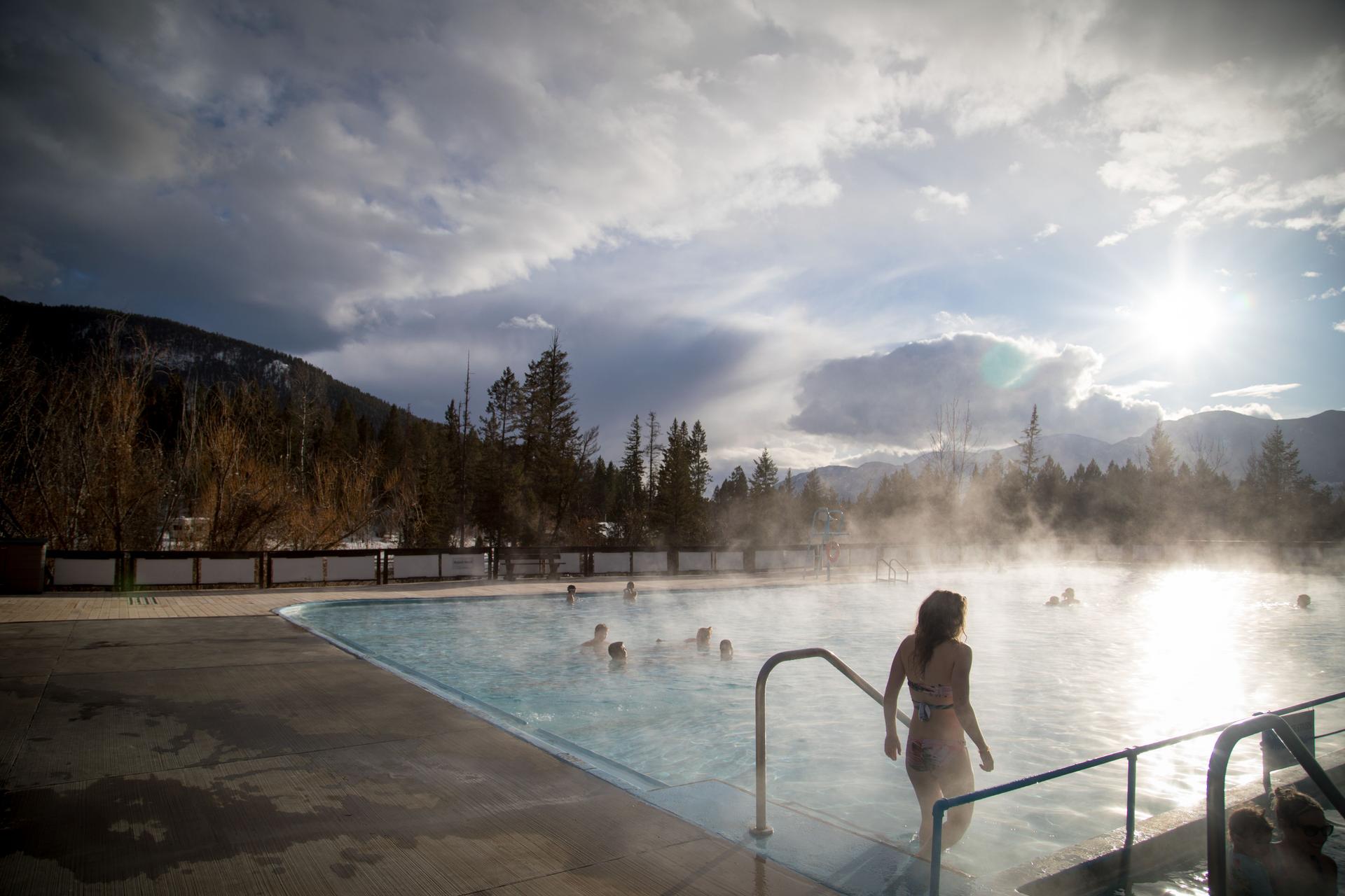 Swimmers soaking in the steaming outdoor mineral pool at Fairmont Hot Springs Resort in winter, with snow-dusted mountains and evergreen trees in the background, Columbia Valley, British Columbia