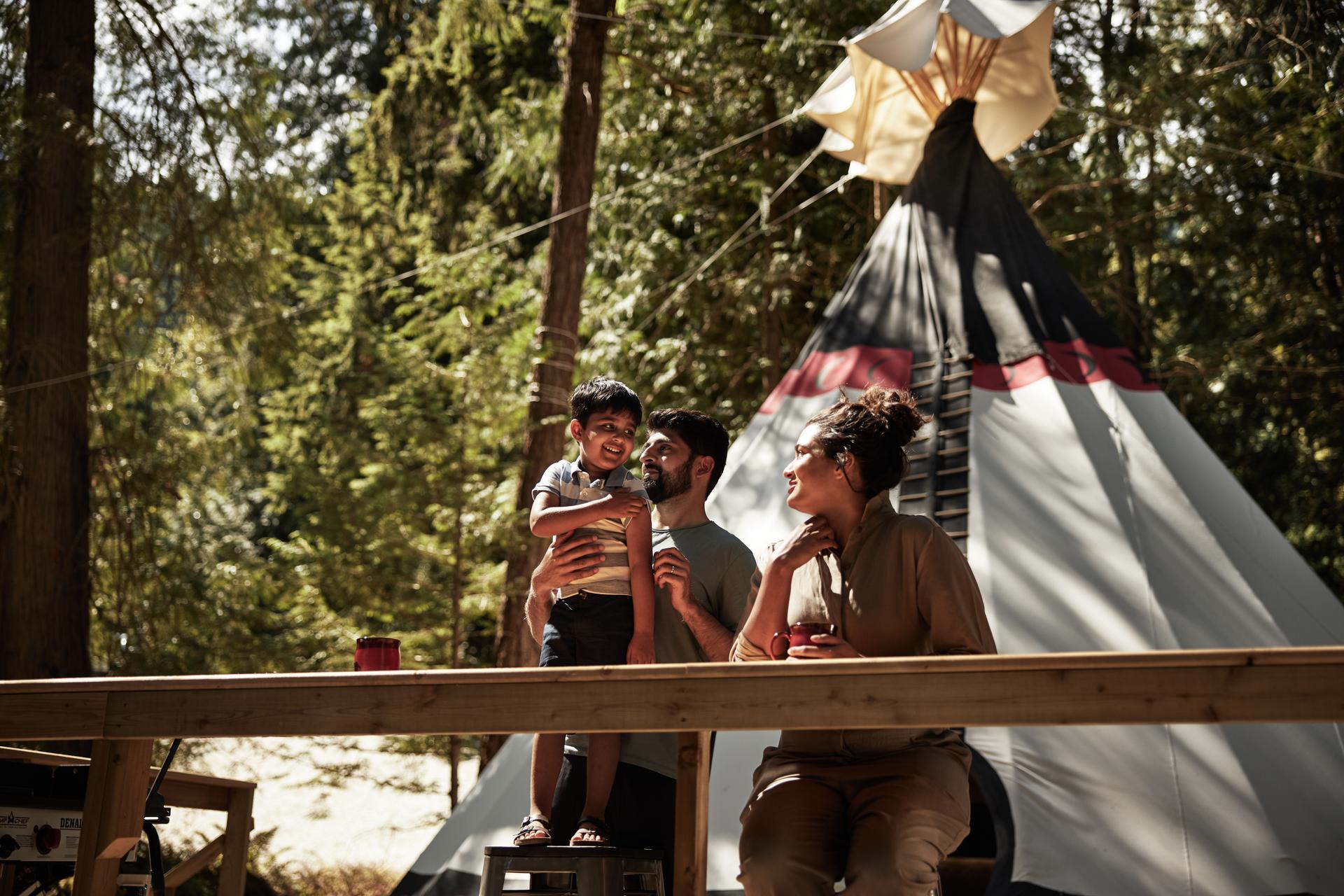 A family with a young child gathers at a table outside a luxury teepee in the forest at Fraser Canyon Teepee Escape in British Columbia