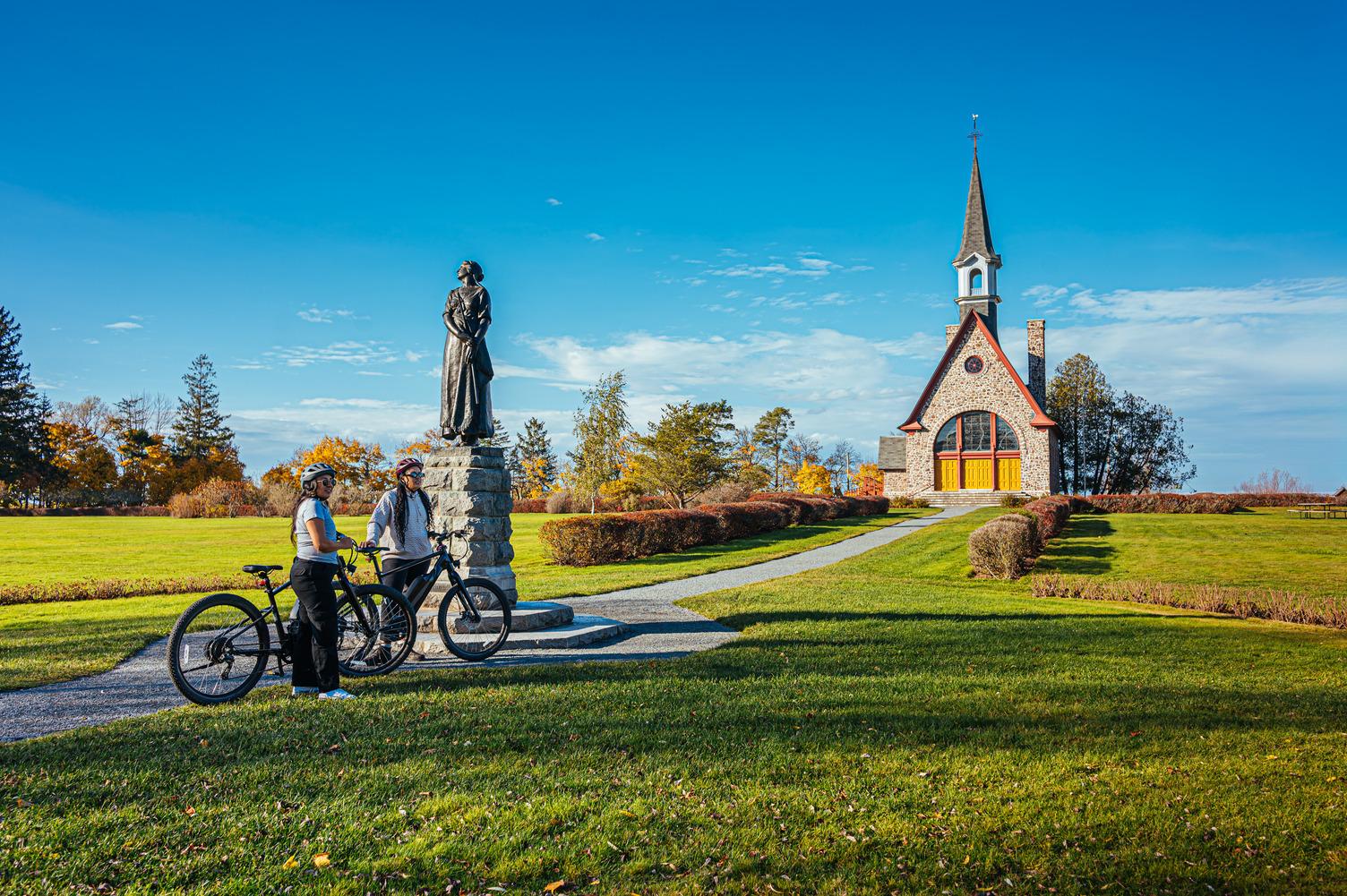 Two cyclists pause beside the Statue of Evangeline at Grand-Pré National Historic Site, with the stone Memorial Church in the background.