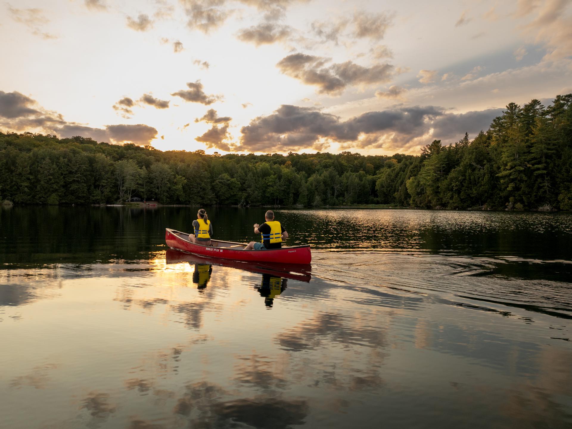 Two paddlers in a red canoe cross a still Haliburton lake as the sun sets behind the treeline