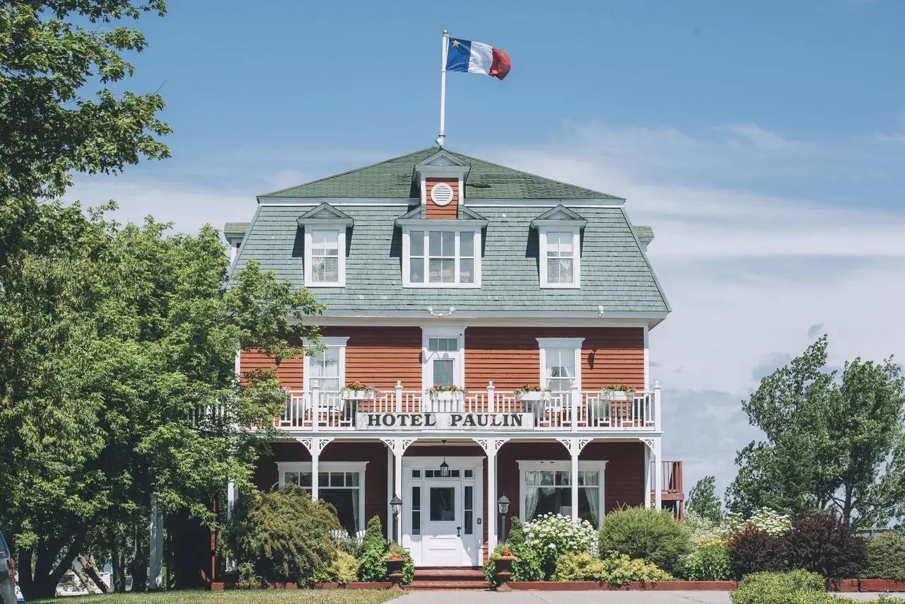 The red-and-white historic exterior of Hotel Paulin in Caraquet, New Brunswick, with an Acadian flag flying above.