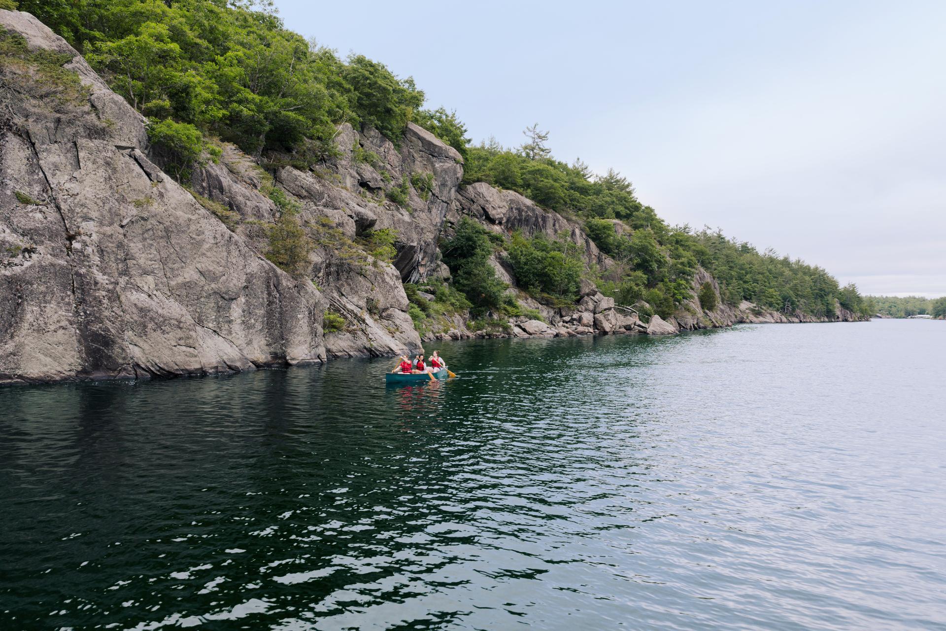 Three canoeists in life jackets paddling below a steep granite cliff face in Kawartha Highlands Provincial Park, Ontario.