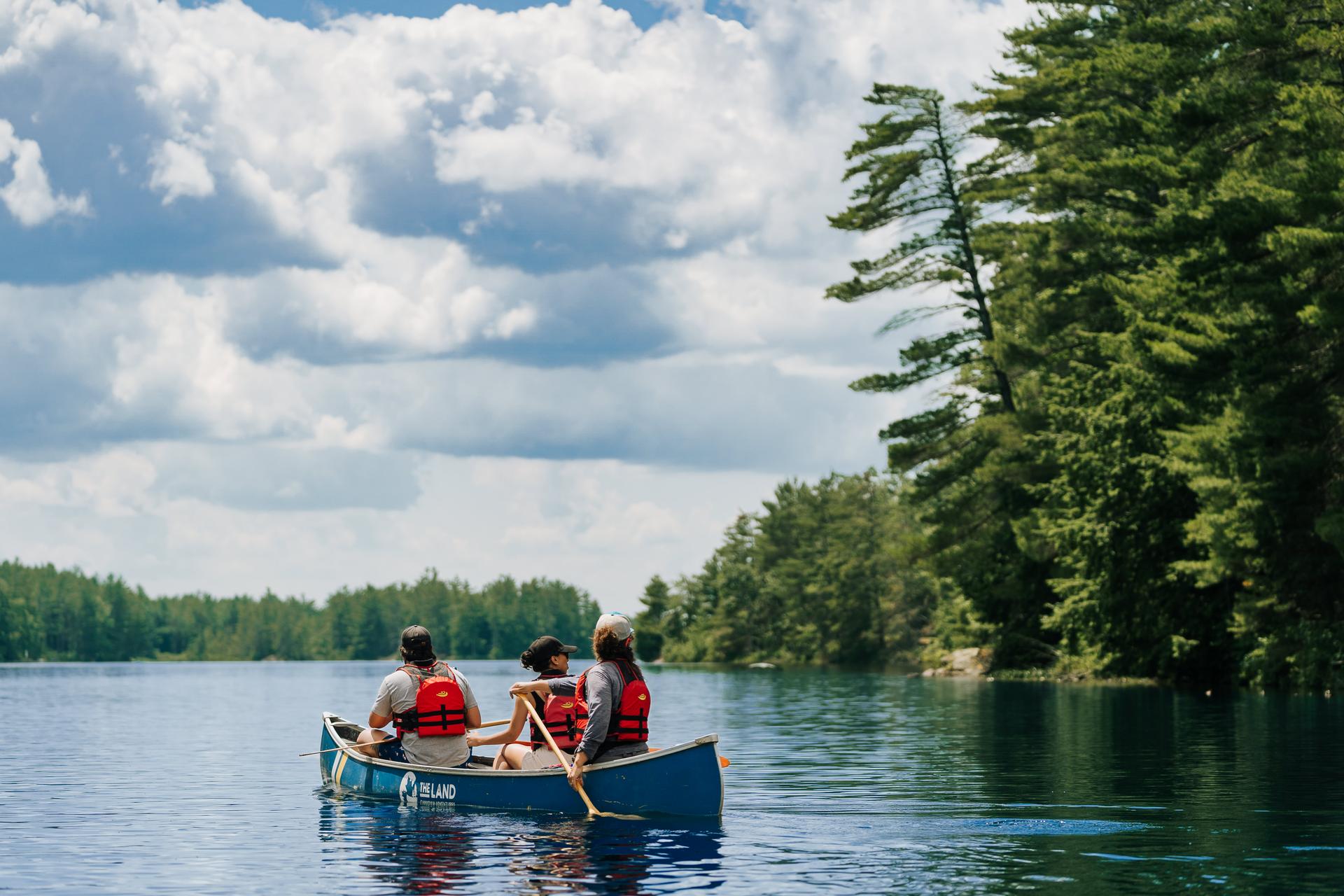Three paddlers in life jackets canoe along a tree-lined Kawartha Lakes shoreline in Ontario