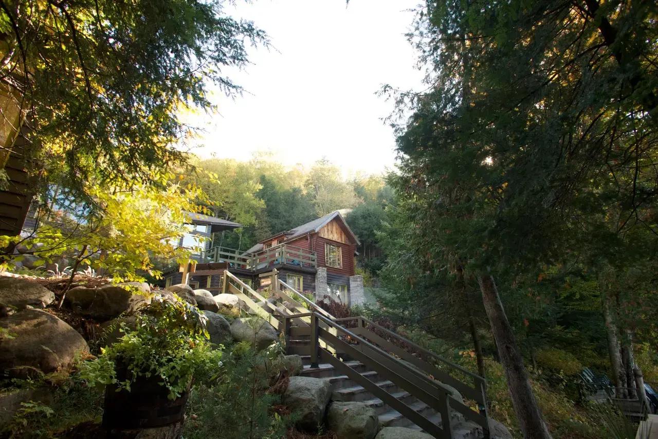A rustic red wood cabin nestled among large boulders and tall evergreen and deciduous trees on a forested hillside at Kayak & Cabana in the Laurentian Mountains of Quebec, Canada, with wooden staircase leading up to the entrance on a sunny autumn day