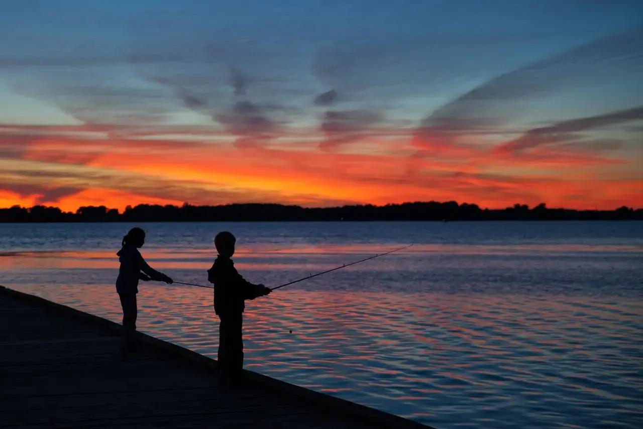 Silhouettes of two children fishing with a rod from a dock at sunset over Lake Erie in Ontario, Canada, with a vivid orange and red sky reflected across the calm water and a dark treeline on the distant shore