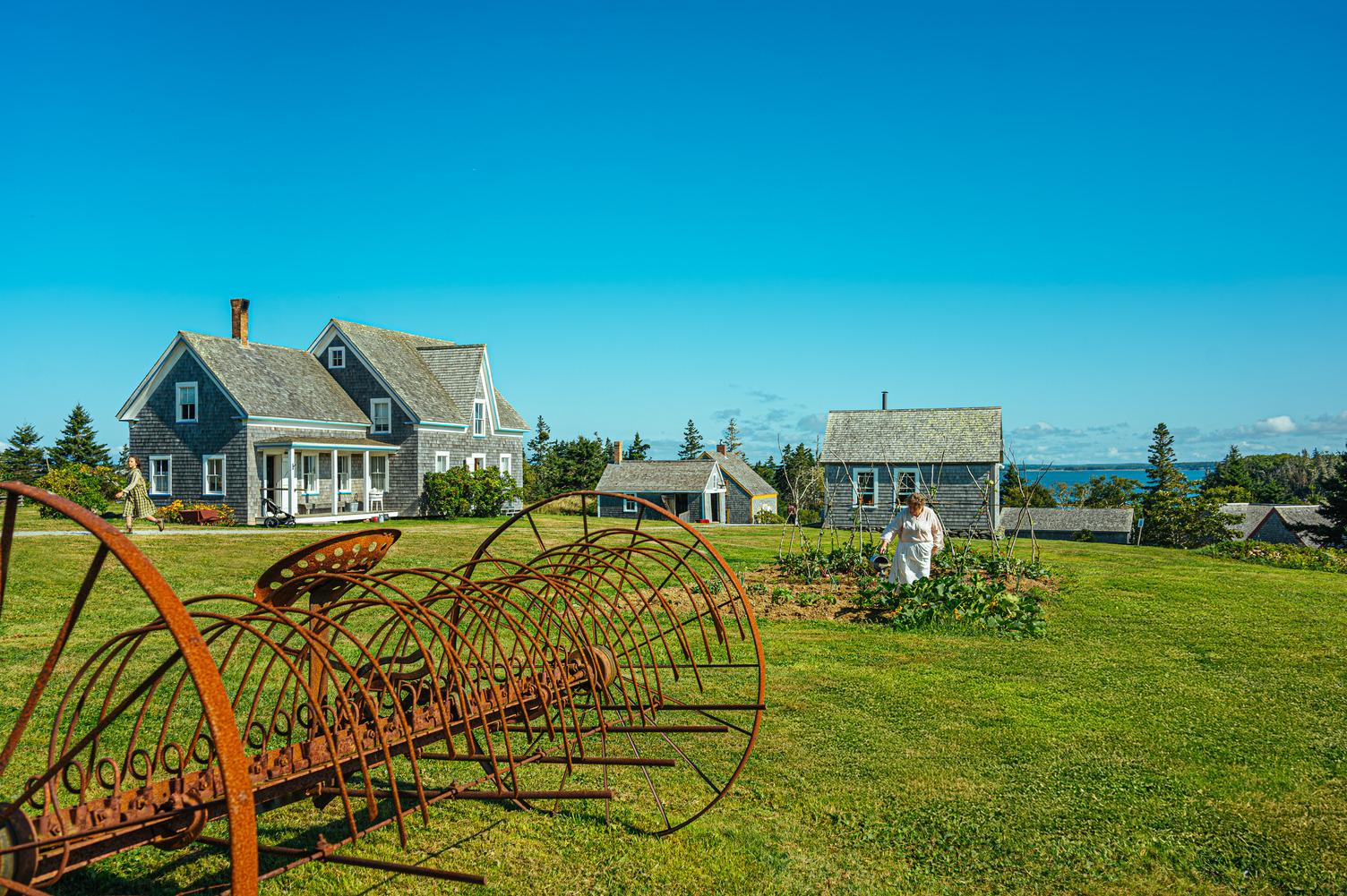 Shingled heritage buildings and a costumed interpreter tending a garden at Le village historique Acadien de la Nouvelle-Écosse, overlooking Pubnico Harbour.