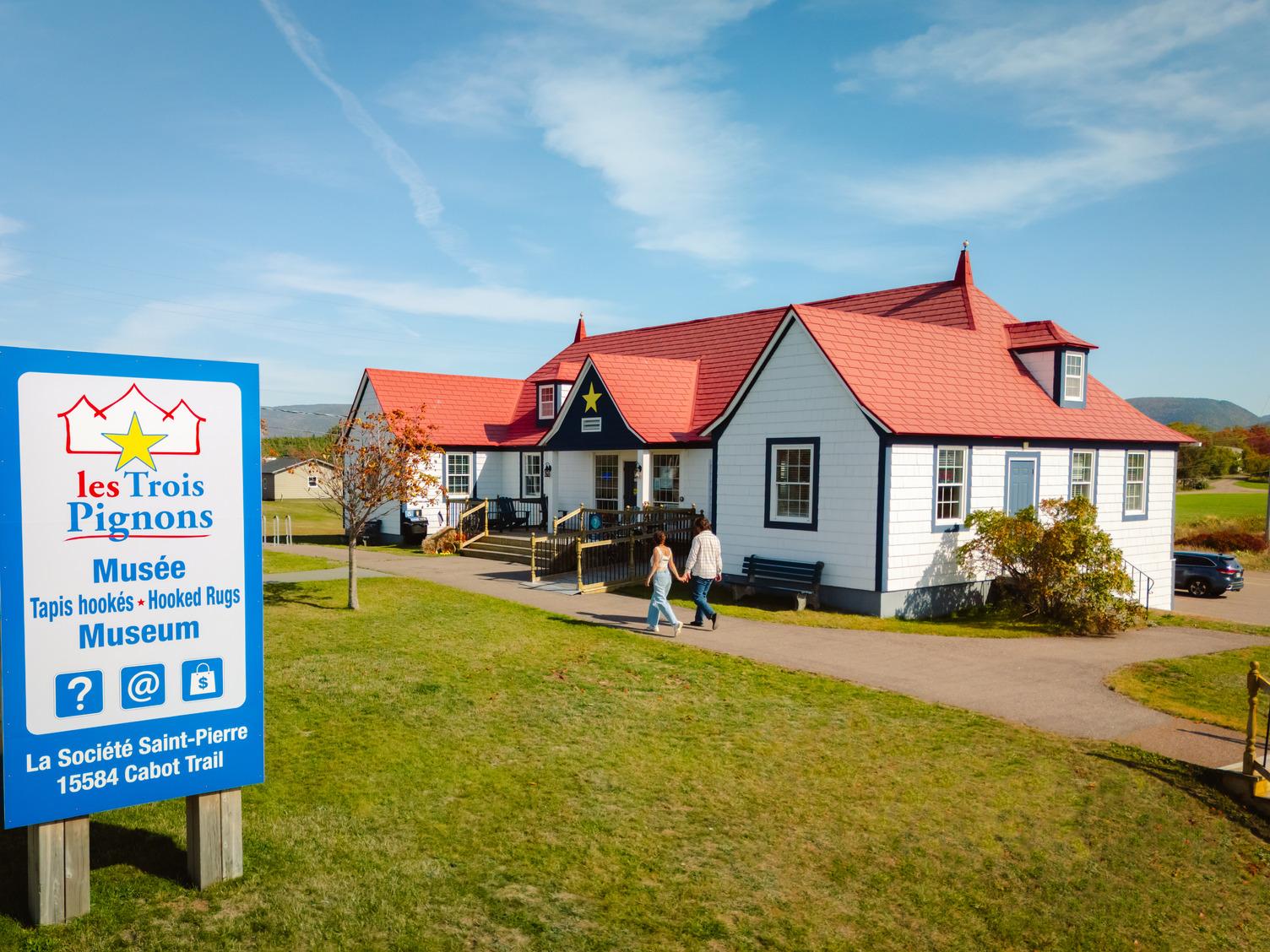 A couple walks toward Les Trois Pignons, the red-roofed Museum of the Hooked Rug and Home Life in Chéticamp, with a signboard in the foreground.